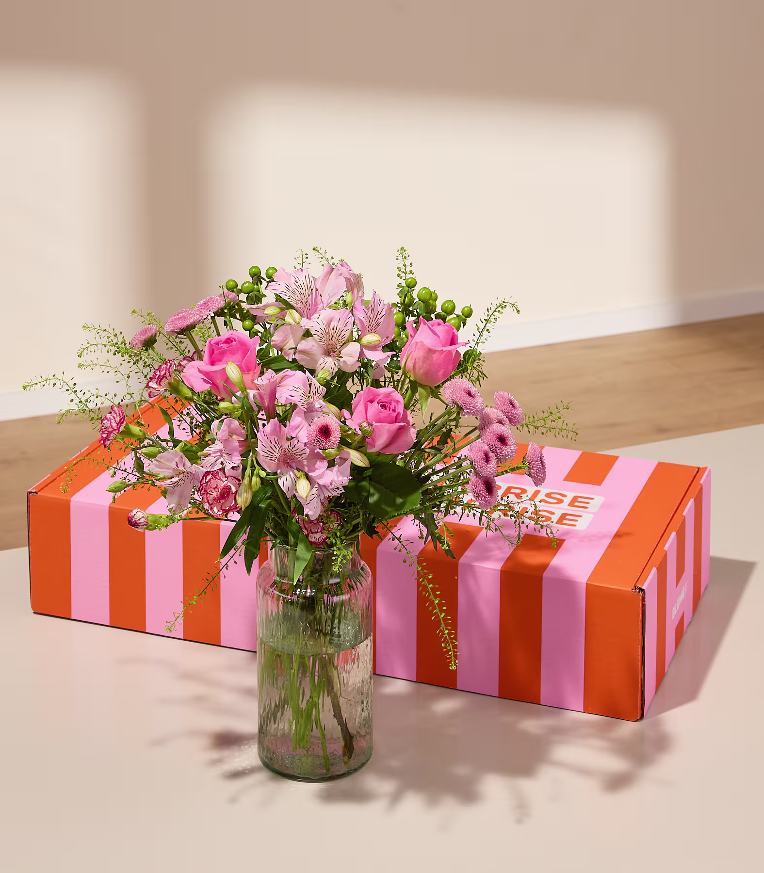 Ein Strauß rosa Blumen in einer Glasvase steht auf einer orange-weiß gestreiften Geschenkbox auf einer Holzoberfläche. (Beschreibungstext KI-generiert)