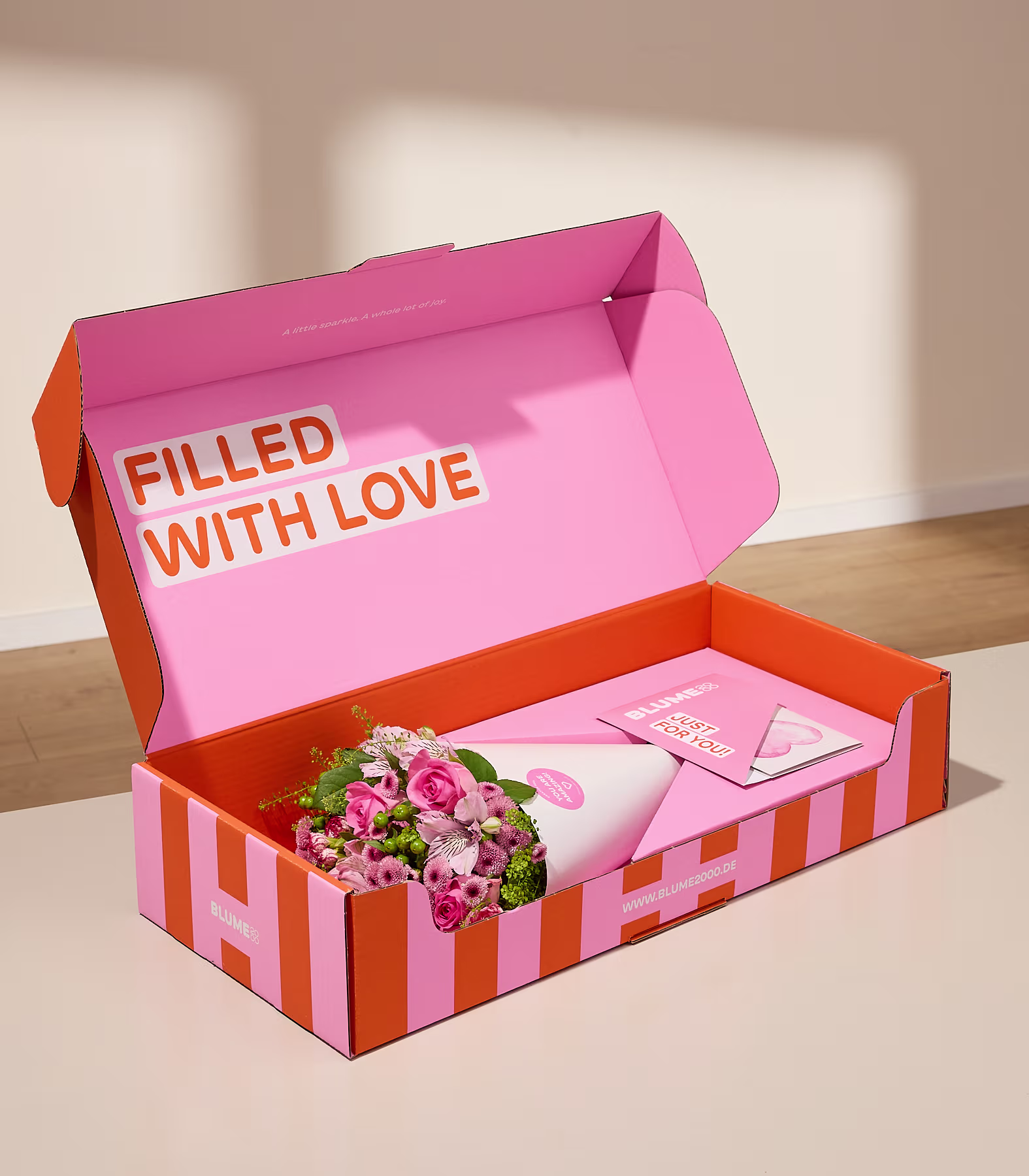 Eine rosa-orange gestreifte Schachtel mit der Aufschrift „VOLL MIT LIEBE“ enthält einen Strauß rosa-weißer Blumen auf einer Holzoberfläche. (Beschreibungstext KI-generiert)