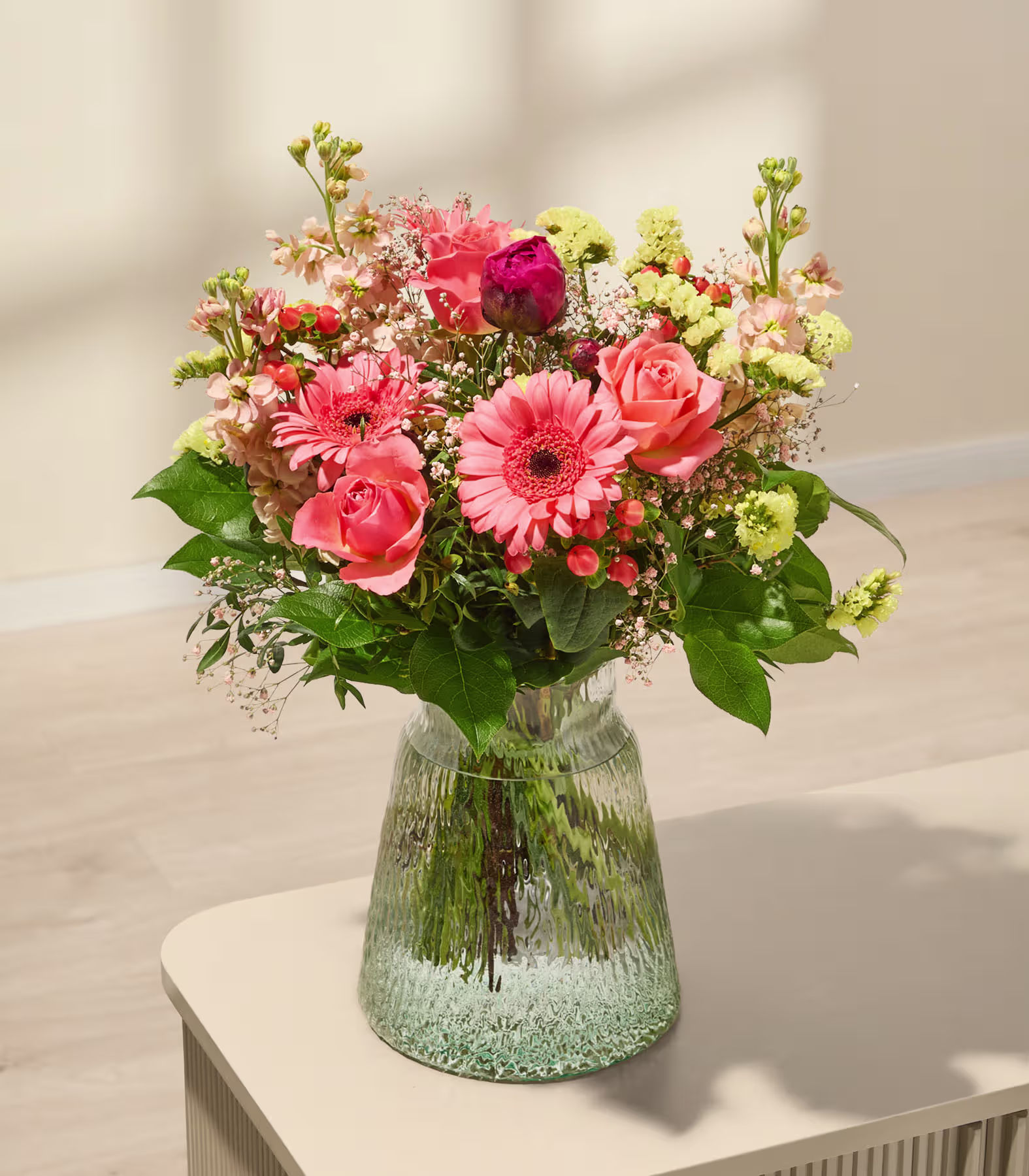 Ein farbenfroher Strauß aus rosa und roten Blumen, darunter Rosen und Gerbera-Gänseblümchen, arrangiert in einer Glasvase auf einer hellen Oberfläche. (Beschreibungstext KI-generiert)