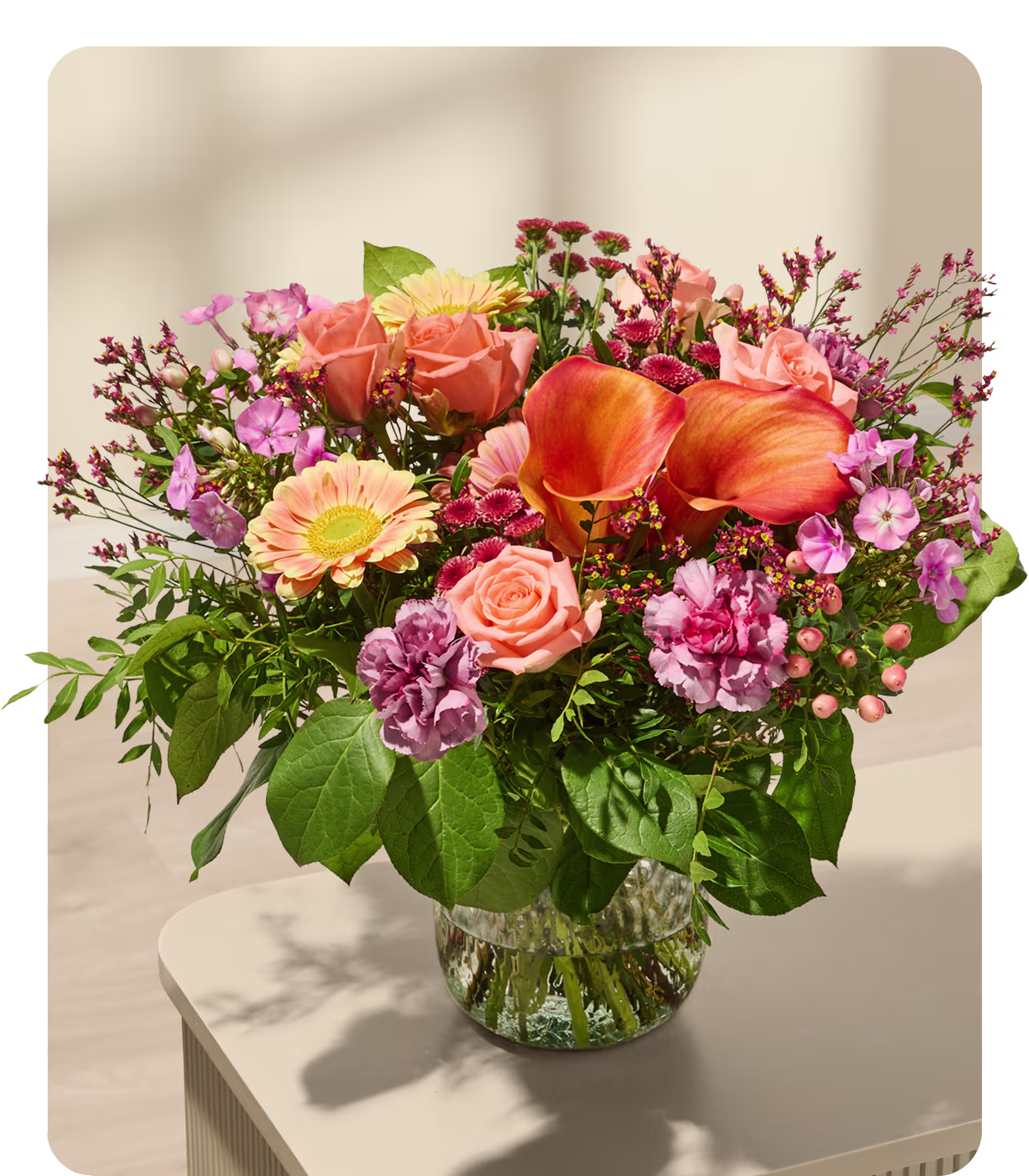 Ein farbenfroher Strauß aus verschiedenen Blumen, darunter Rosen, Gerbera und Nelken, arrangiert in einer Glasvase vor einem neutralen Hintergrund. (Beschreibungstext KI-generiert)