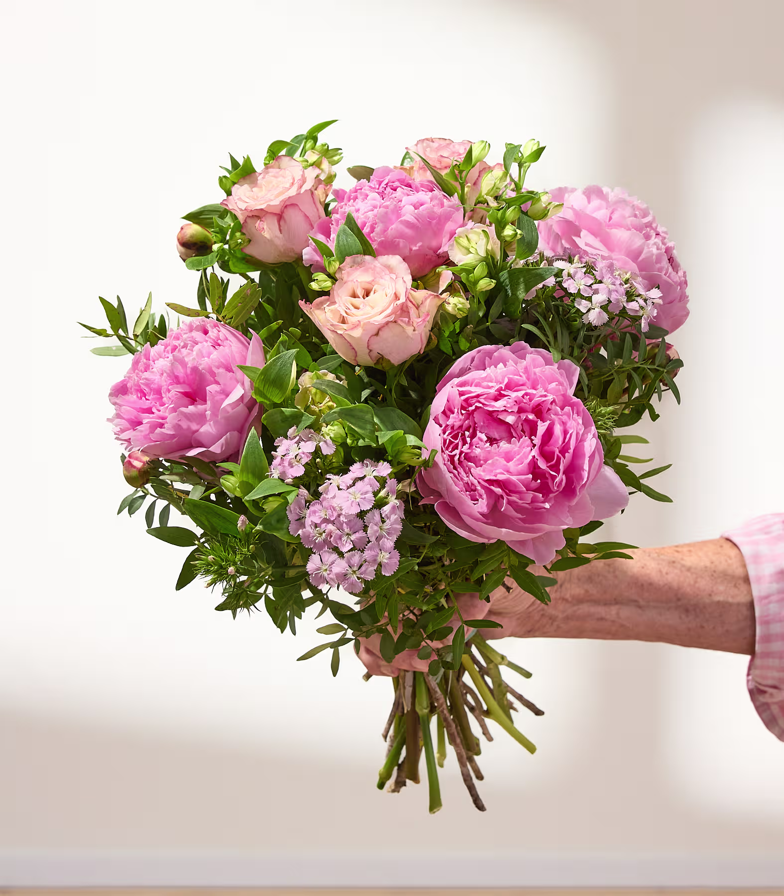 Ein leuchtender Strauß aus rosa Pfingstrosen, weißen Blumen und Grünpflanzen wird in den Händen einer Person gehalten, die vor einem schlichten Hintergrund einen blauen Anzug trägt. (Beschreibungstext KI-generiert)