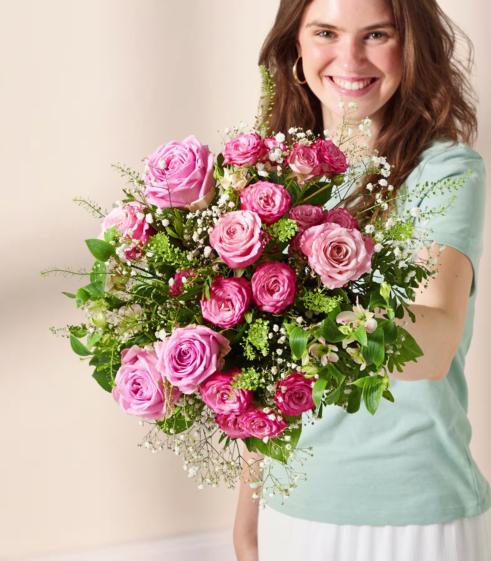 Eine junge Frau mit einem warmen Lächeln hält vor einem neutralen Hintergrund einen leuchtenden Strauß aus rosa Rosen und anderen Blumen. (Beschreibungstext KI-generiert)