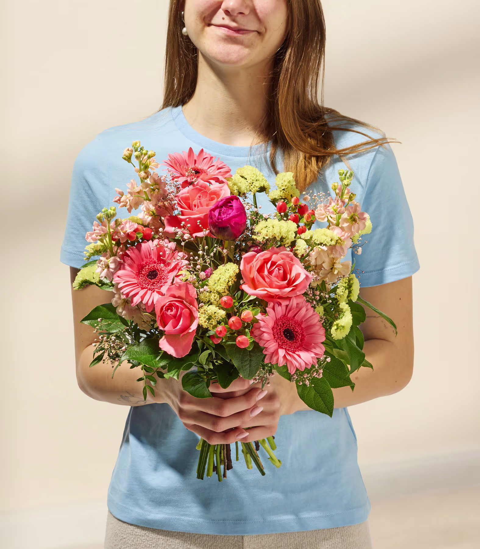 Eine junge Frau in einem blauen Hemd hält einen farbenfrohen Strauß rosa und gelber Blumen vor einem schlichten beigen Hintergrund. (Beschreibungstext KI-generiert)