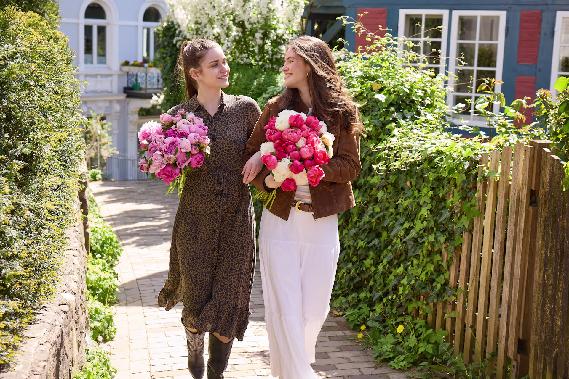 Zwei junge Frauen in legerer Kleidung stehen in einem üppigen, farbenfrohen Garten und halten große Sträuße leuchtender Blumen. (Beschreibungstext KI-generiert)