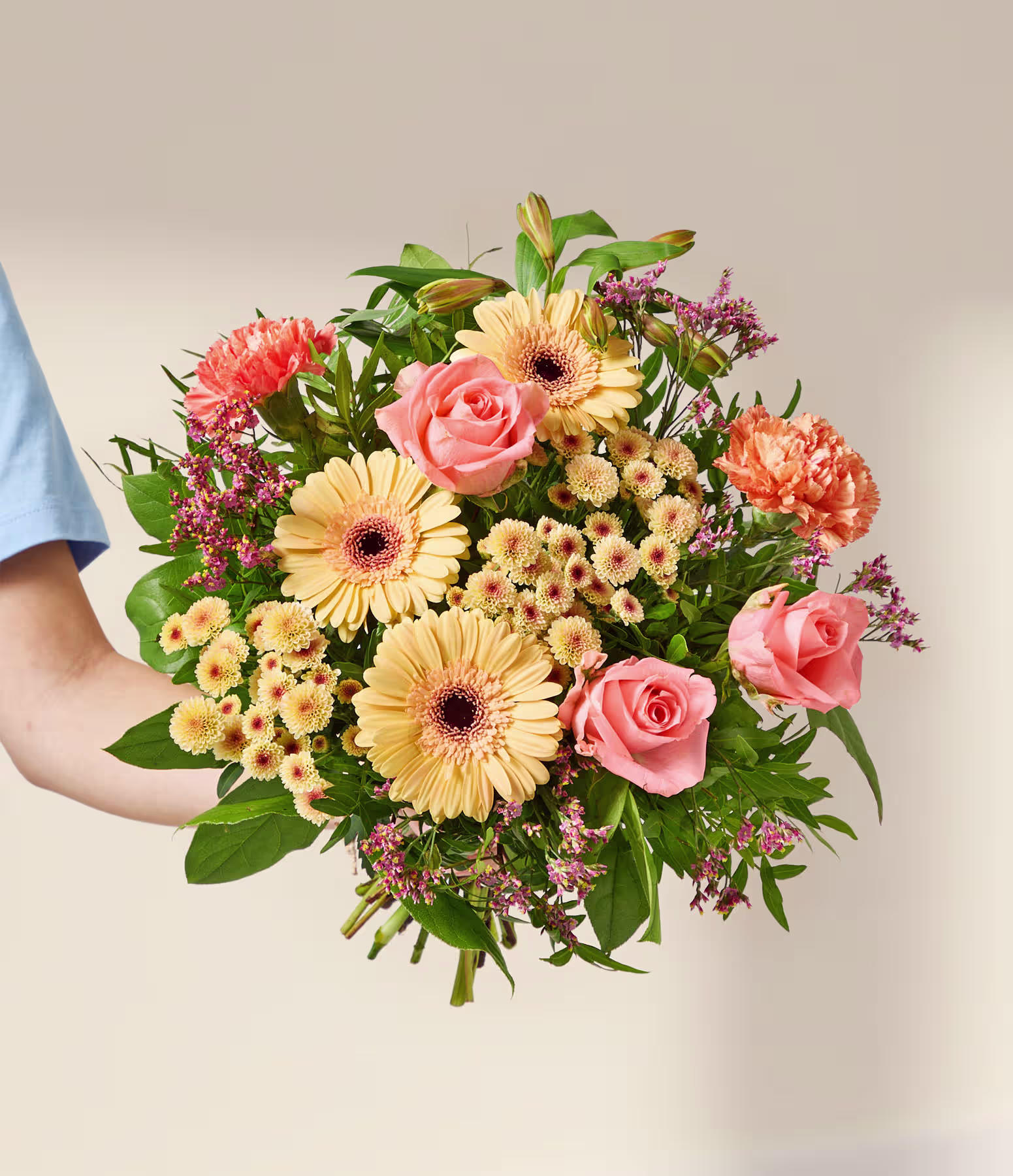 Eine Hand hält einen leuchtenden Strauß aus rosa Rosen, orangefarbenen Nelken und gelben Gerbera vor einem sanften, neutralen Hintergrund. (Beschreibungstext KI-generiert)