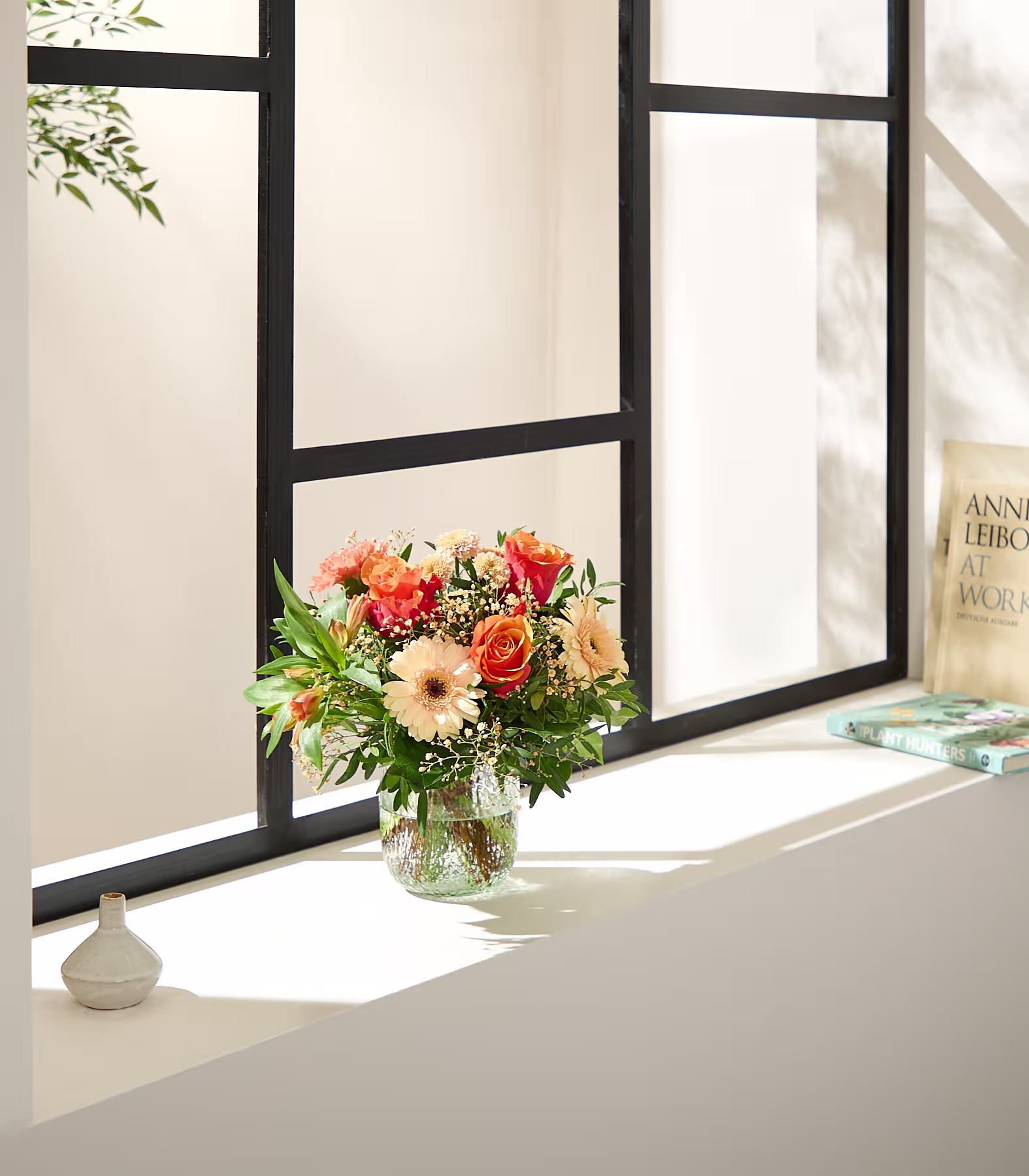 Auf dem Fensterbrett steht ein leuchtender Strauß orangefarbener und rosafarbener Blumen in einer Glasvase, rechts daneben ein Buch und eine kleine Vase, und im Hintergrund ein Fenster mit Blick ins Grüne. (Beschreibungstext KI-generiert)