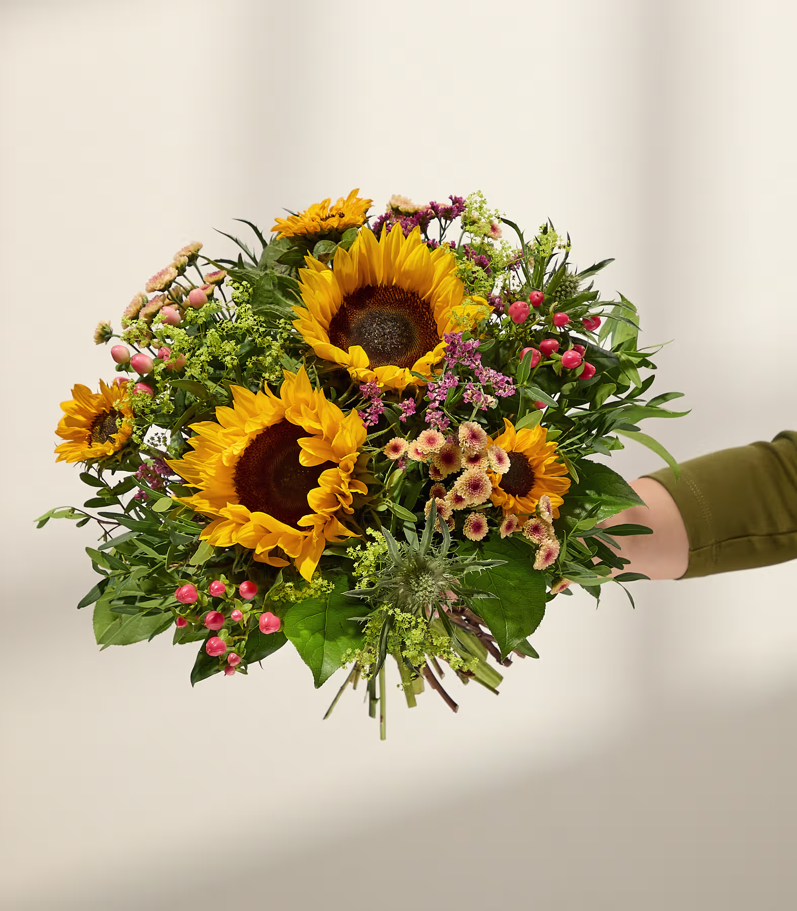 Eine Hand hält einen leuchtenden Strauß Sonnenblumen und kleineren rosa und gelben Blüten vor einem sanft beleuchteten, neutralen Hintergrund. (Beschreibungstext KI-generiert)