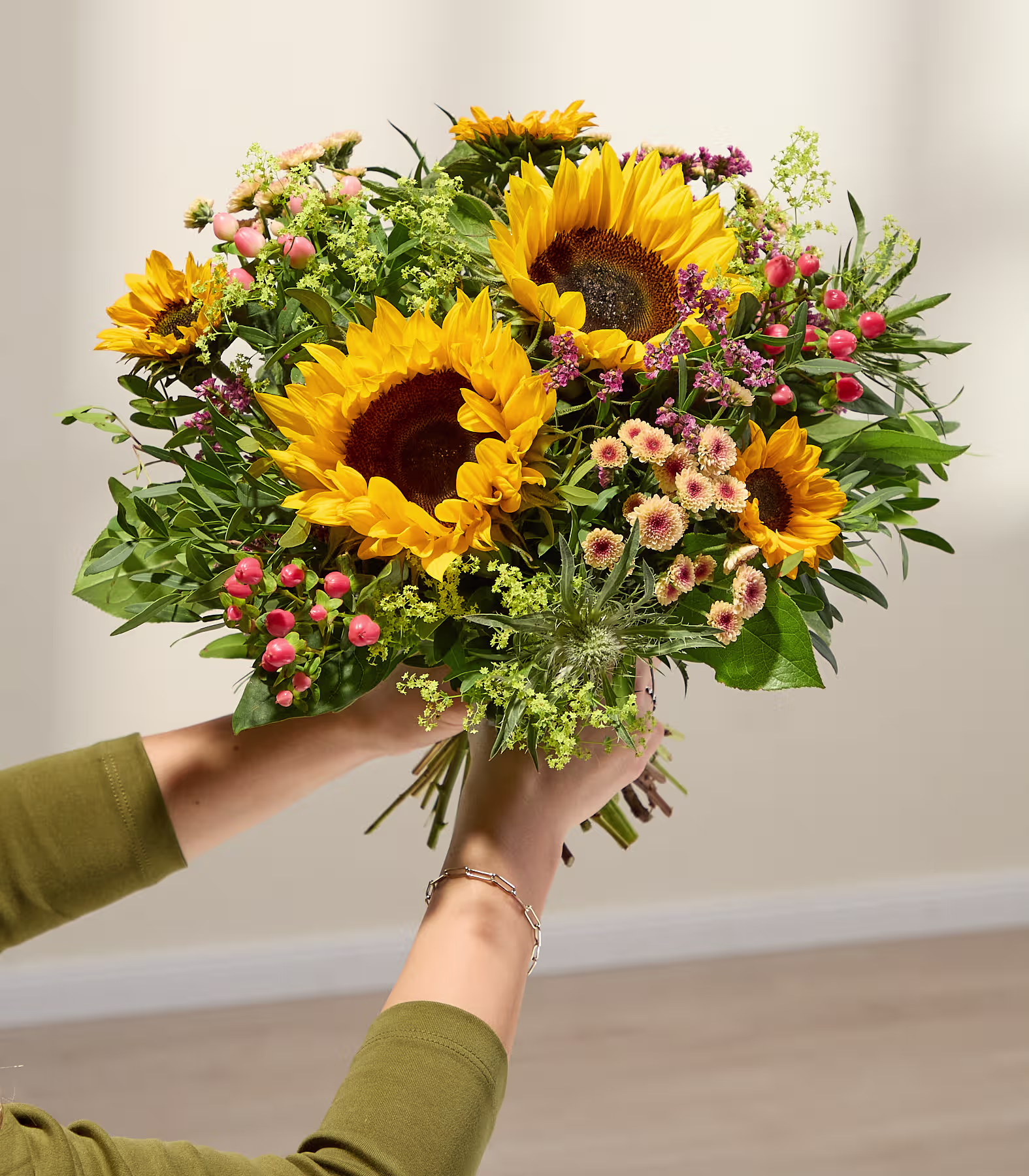 Im Vordergrund hält ein leuchtender Strauß Sonnenblumen und anderer Wildblumen die Hände vor einem sanft beleuchteten, neutralen Hintergrund. (Beschreibungstext KI-generiert)