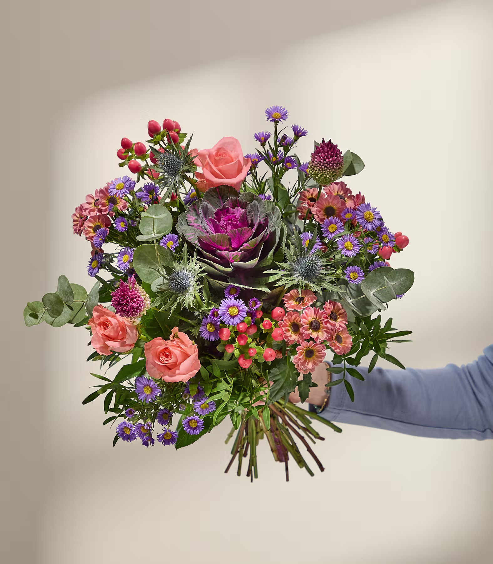 Eine Hand in einem blauen Ärmel hält einen leuchtenden Strauß aus Rosen, Disteln und Zierkohl vor einem sanft beleuchteten, neutralen Hintergrund. (Beschreibungstext KI-generiert)