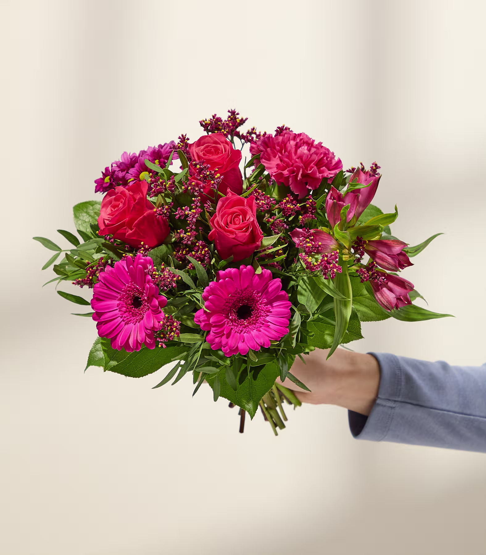 Eine Hand in einem blauen Ärmel hält einen leuchtenden Strauß rosa und roter Blumen vor einem sanft beleuchteten, neutralen Hintergrund. (Beschreibungstext KI-generiert)