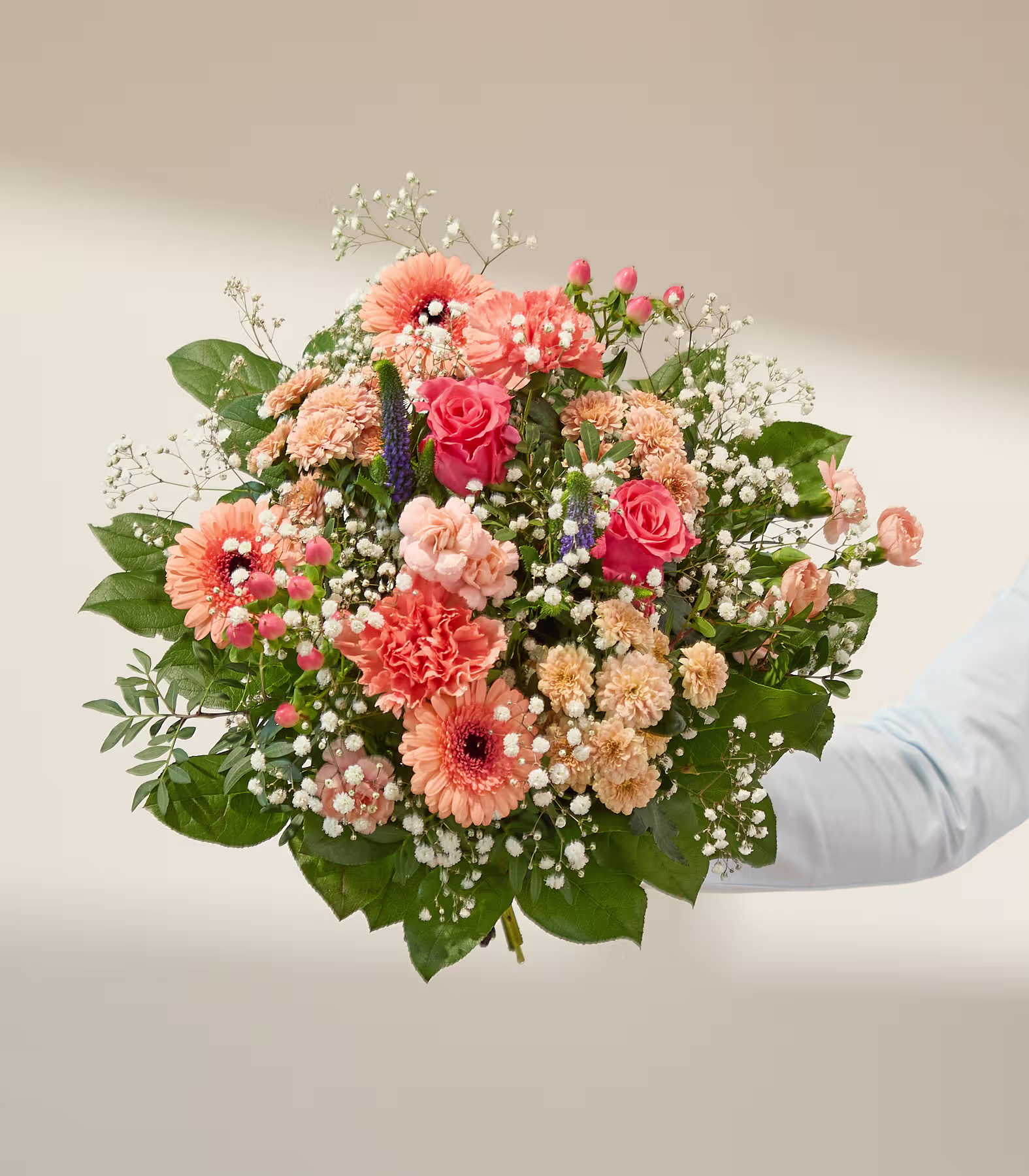 Ein lebhafter Blumenstrauß in Pfirsich- und Rosatönen, akzentuiert mit Schleierkraut und Grünzeug, hebt sich von einem sanft beleuchteten, neutralen Hintergrund ab. (Beschreibungstext KI-generiert)