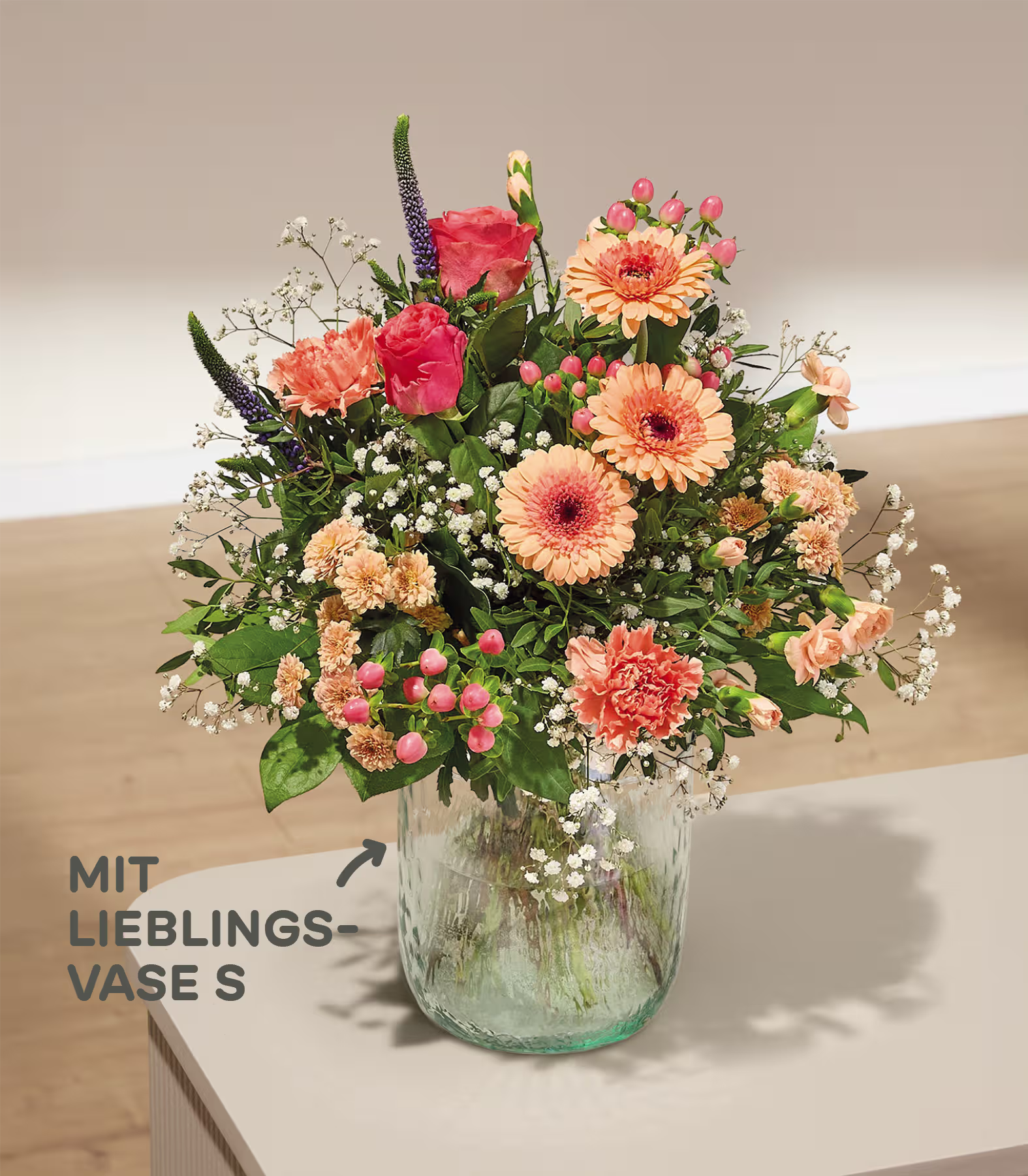 Ein leuchtender Strauß rosa und orangefarbener Blumen füllt eine strukturierte grüne Vase auf hellem Untergrund; im Hintergrund sind Wand und Boden sanft verschwommen zu sehen. (Beschreibungstext KI-generiert)