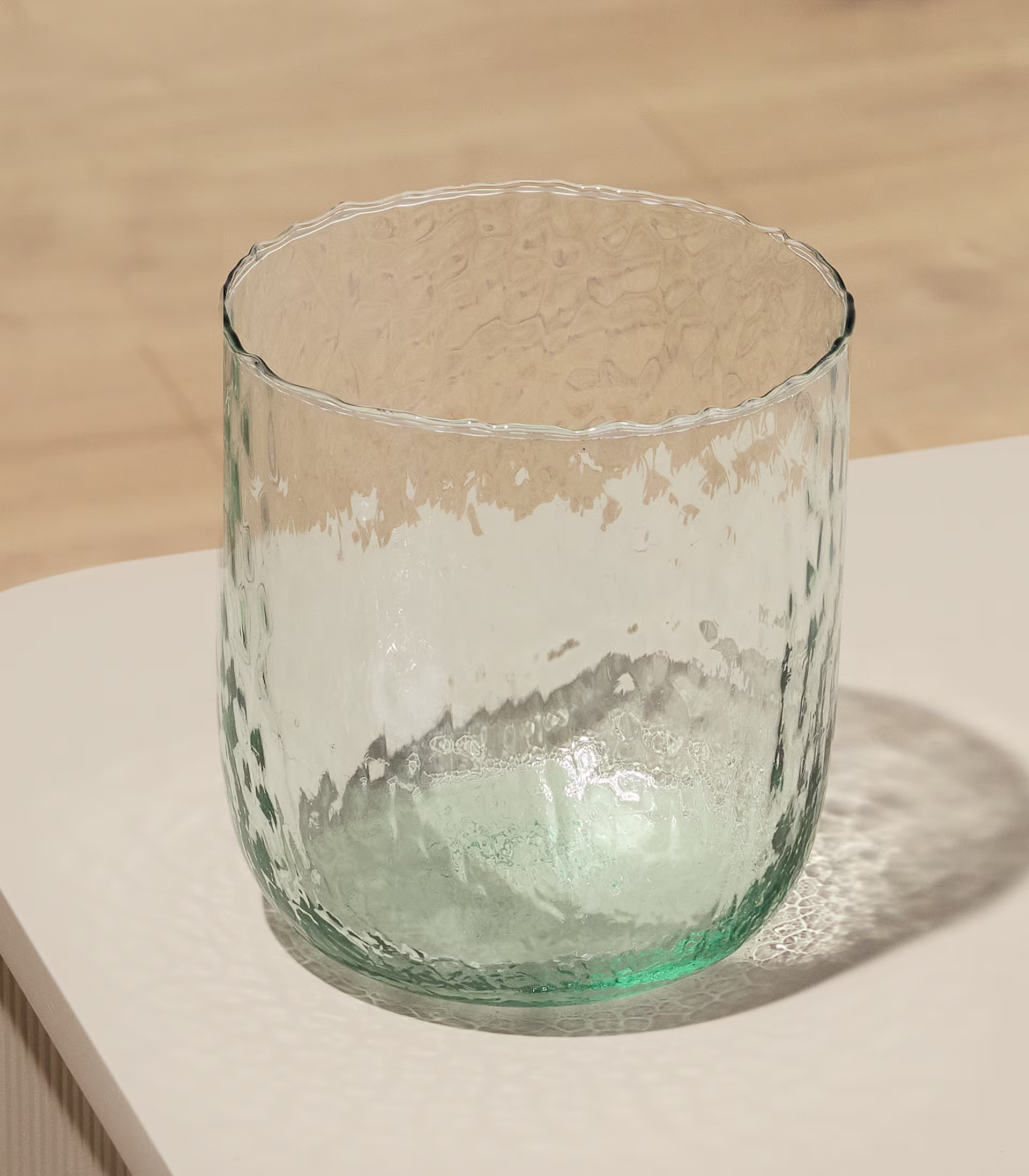 Ein durchsichtiges Glas mit einer strukturierten, welligen Oberfläche steht auf einem Holztisch vor einem schlichten, hellen Hintergrund. (Beschreibungstext KI-generiert)