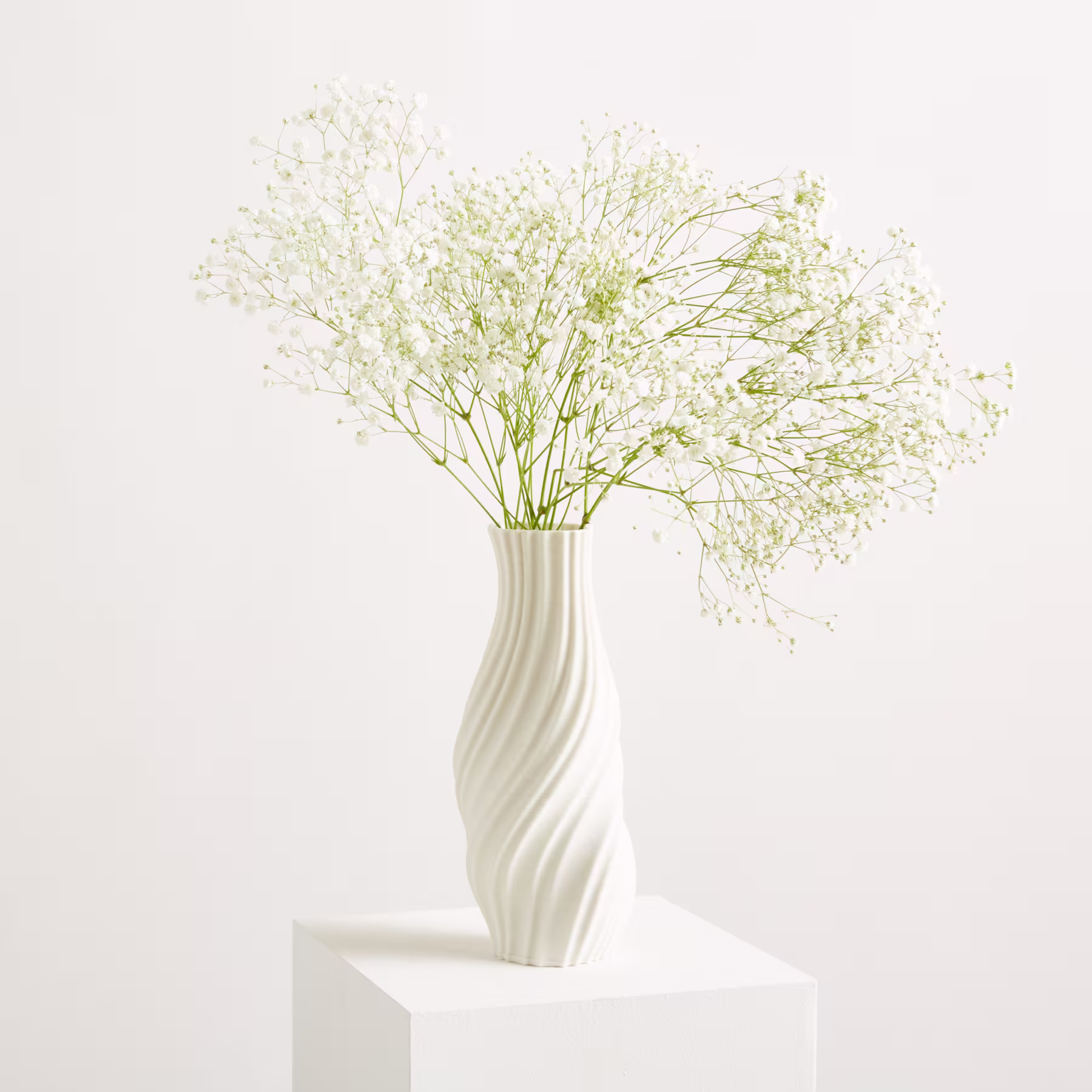 Eine weiße Vase mit zarten weißen Blüten steht auf einem weißen Sockel vor einem schlichten weißen Hintergrund. (Beschreibungstext KI-generiert)