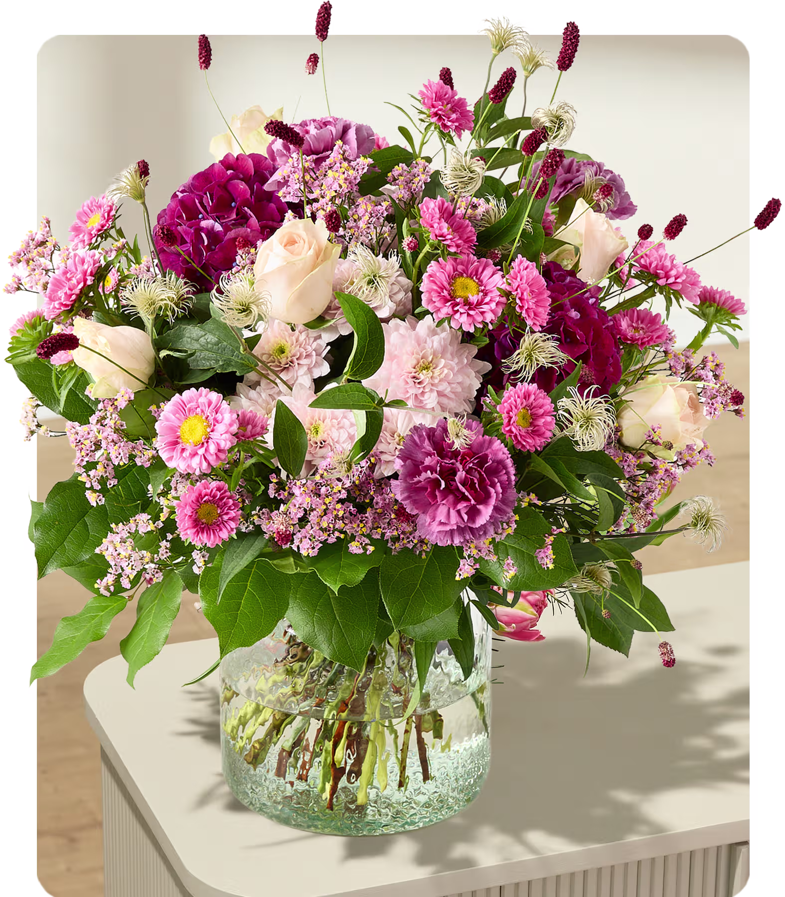 Ein üppiger Strauß rosa und weißer Blumen quillt aus einer durchsichtigen Glasvase auf einer hellen Oberfläche vor einem sanft verschwommenen Hintergrund. (Beschreibungstext KI-generiert)