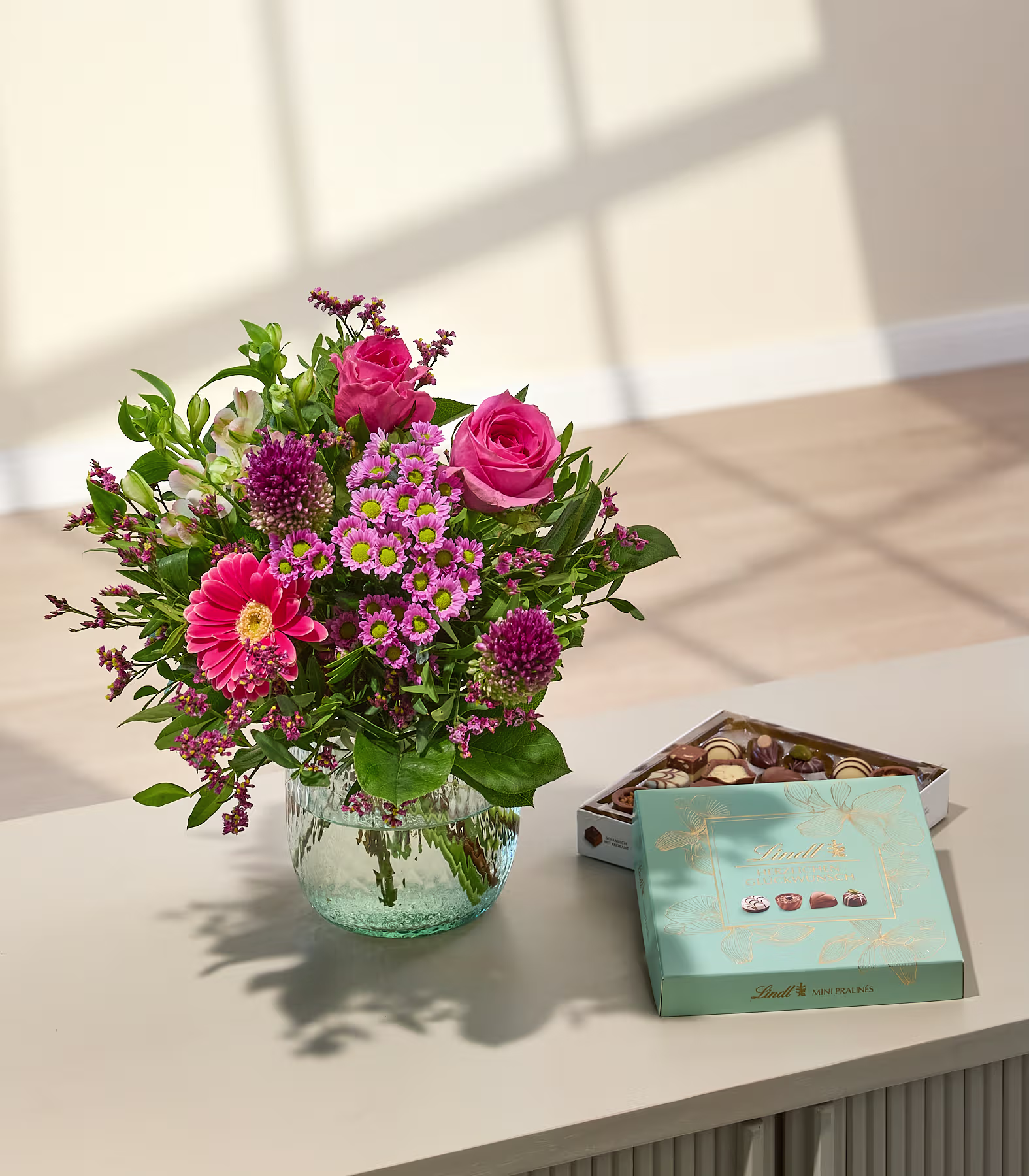 Auf einem Tisch steht ein farbenfroher Strauß rosa und lila Blumen in einer Glasvase neben einer Schachtel Pralinen; im Hintergrund ist eine sanft beleuchtete Wand zu sehen. (Beschreibungstext KI-generiert)