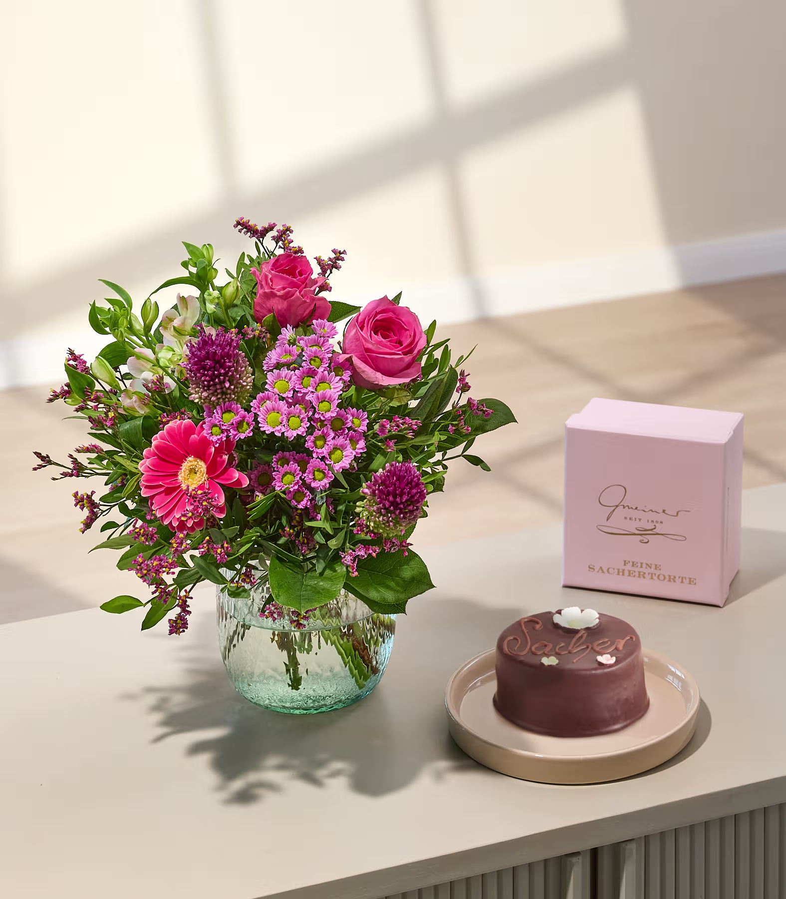 Ein leuchtender Strauß rosa Blumen steht in einer Glasvase neben einem Schokoladenkuchen und einer rosa Geschenkbox auf einer hellen Oberfläche, im Hintergrund ist eine sanft beleuchtete Wand zu sehen. (Beschreibungstext KI-generiert)