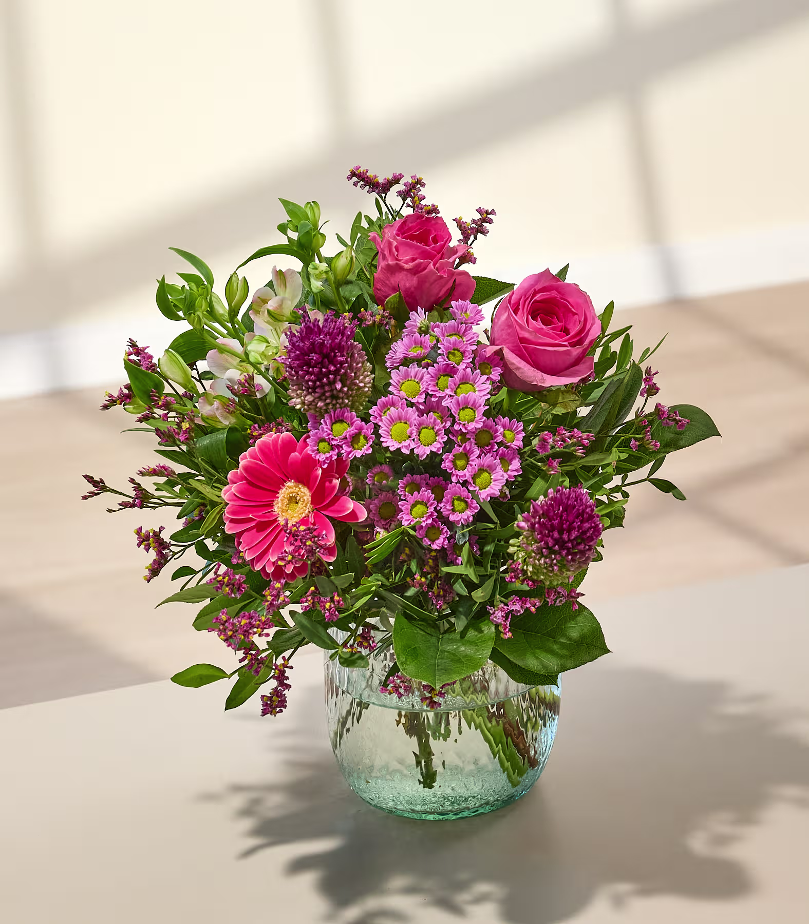 Ein leuchtender Strauß rosa und violetter Blumen in einer durchsichtigen Glasvase steht auf einer hellen Oberfläche, während sanftes Sonnenlicht Schatten auf eine helle Wand im Hintergrund wirft. (Beschreibungstext KI-generiert)
