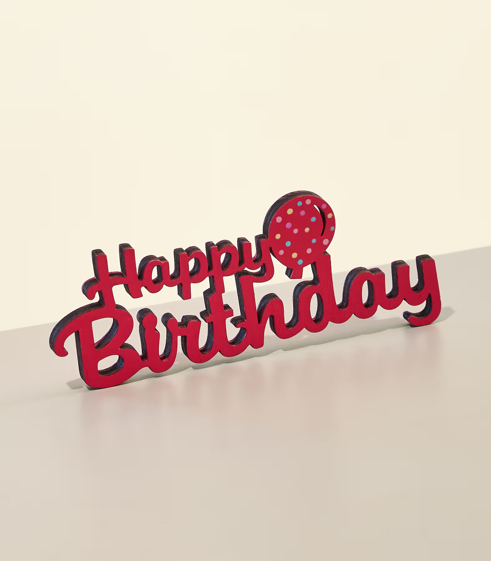 Ein roter „Happy Birthday“-Kuchenaufsatz mit einem gepunkteten Ballon steht auf einer hellen Oberfläche vor einem sanften, cremeweißen Hintergrund. (Beschreibungstext KI-generiert)