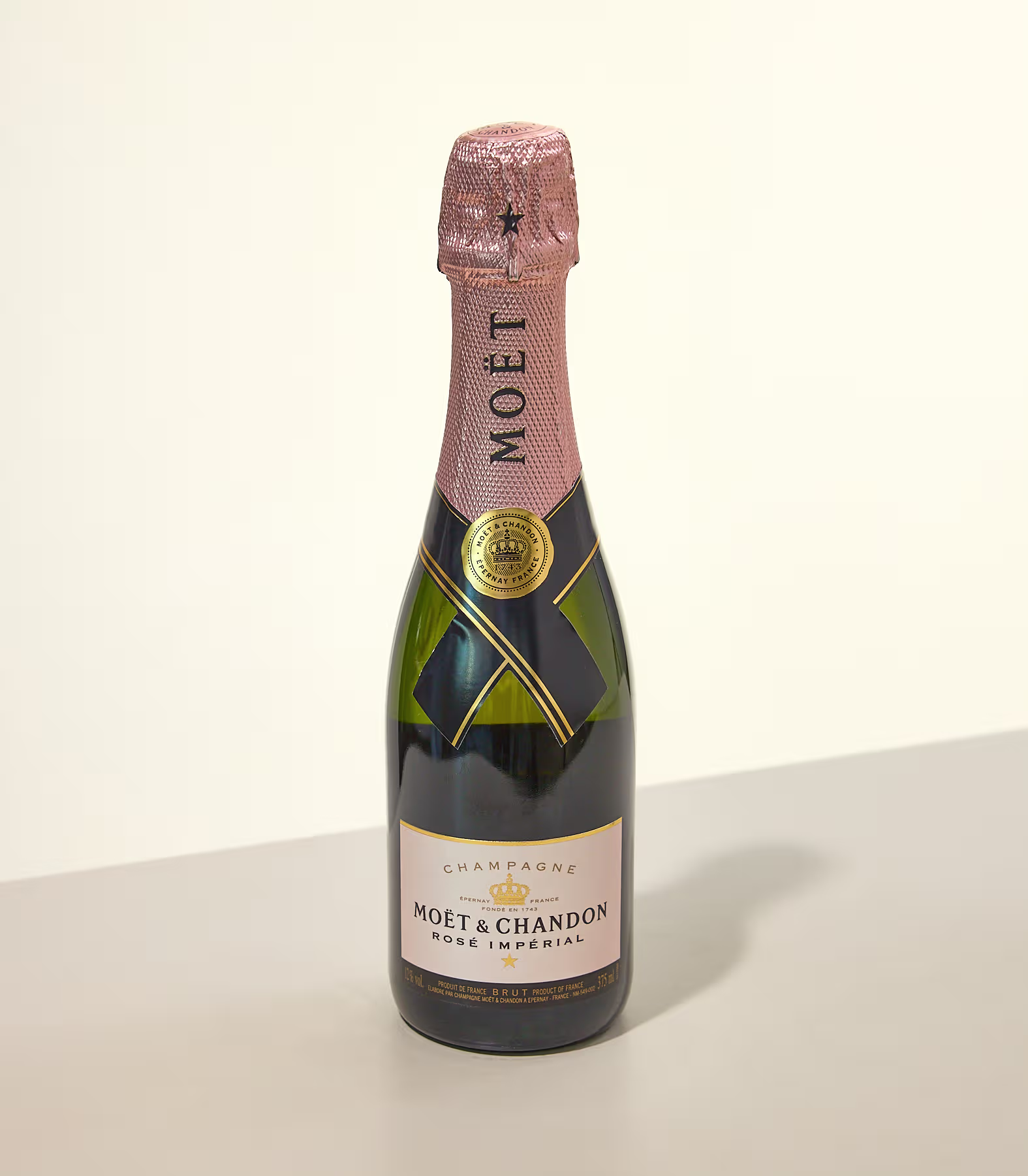 Eine kleine Flasche Moët & Chandon Rosé Imperial Champagner steht auf einer hellgrauen Oberfläche vor einem schlichten, cremeweißen Hintergrund. (Beschreibungstext KI-generiert)