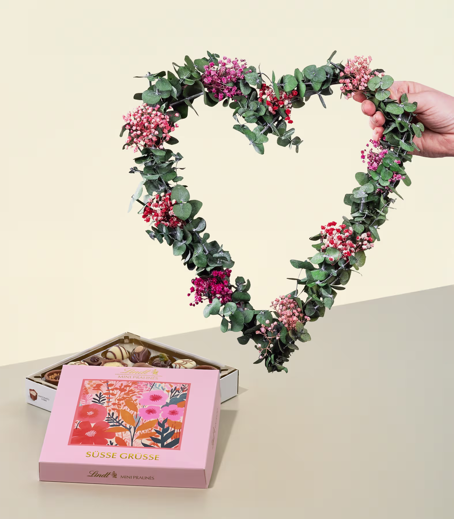 Im Vordergrund wird ein herzförmiger Blumenkranz gehalten, im Hintergrund steht eine Schachtel mit verschiedenen Pralinen. (Beschreibungstext KI-generiert)