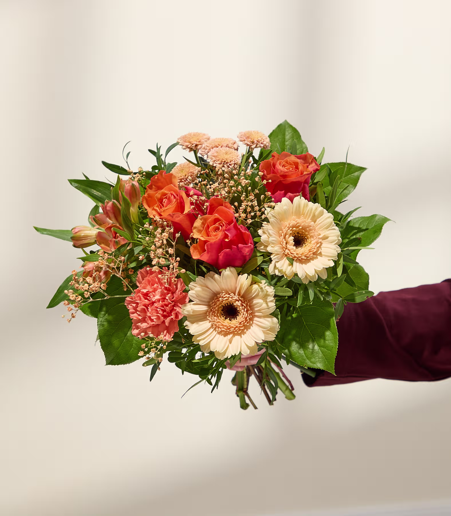Eine Hand in einem bordeauxroten Ärmel hält einen leuchtenden Strauß orangefarbener und pfirsichfarbener Blüten vor einem sanft beleuchteten, neutralen Hintergrund. (Beschreibungstext KI-generiert)