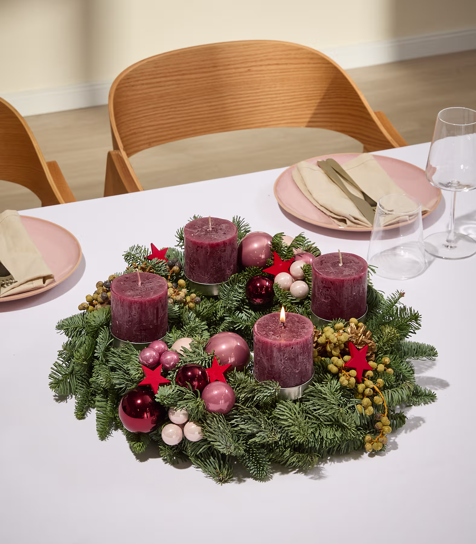 Auf einer weißen Tischdecke steht ein festlicher Adventskranz mit vier brennenden Kerzen, umgeben von Tellern und Gläsern; im sanft beleuchteten Hintergrund sind Holzstühle zu erkennen. (Beschreibungstext KI-generiert)