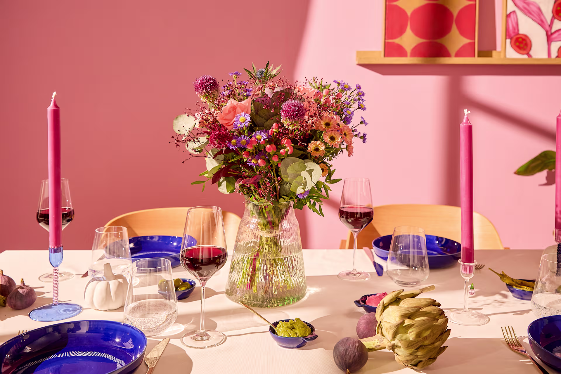Ein für eine Mahlzeit gedeckter Tisch mit einem farbenfrohen Blumenarrangement in der Mitte und zwei hohen rosa Kerzen im Vordergrund, vor einer zartrosa Wand mit dekorativen Regalen im Hintergrund. (Beschreibungstext KI-generiert)