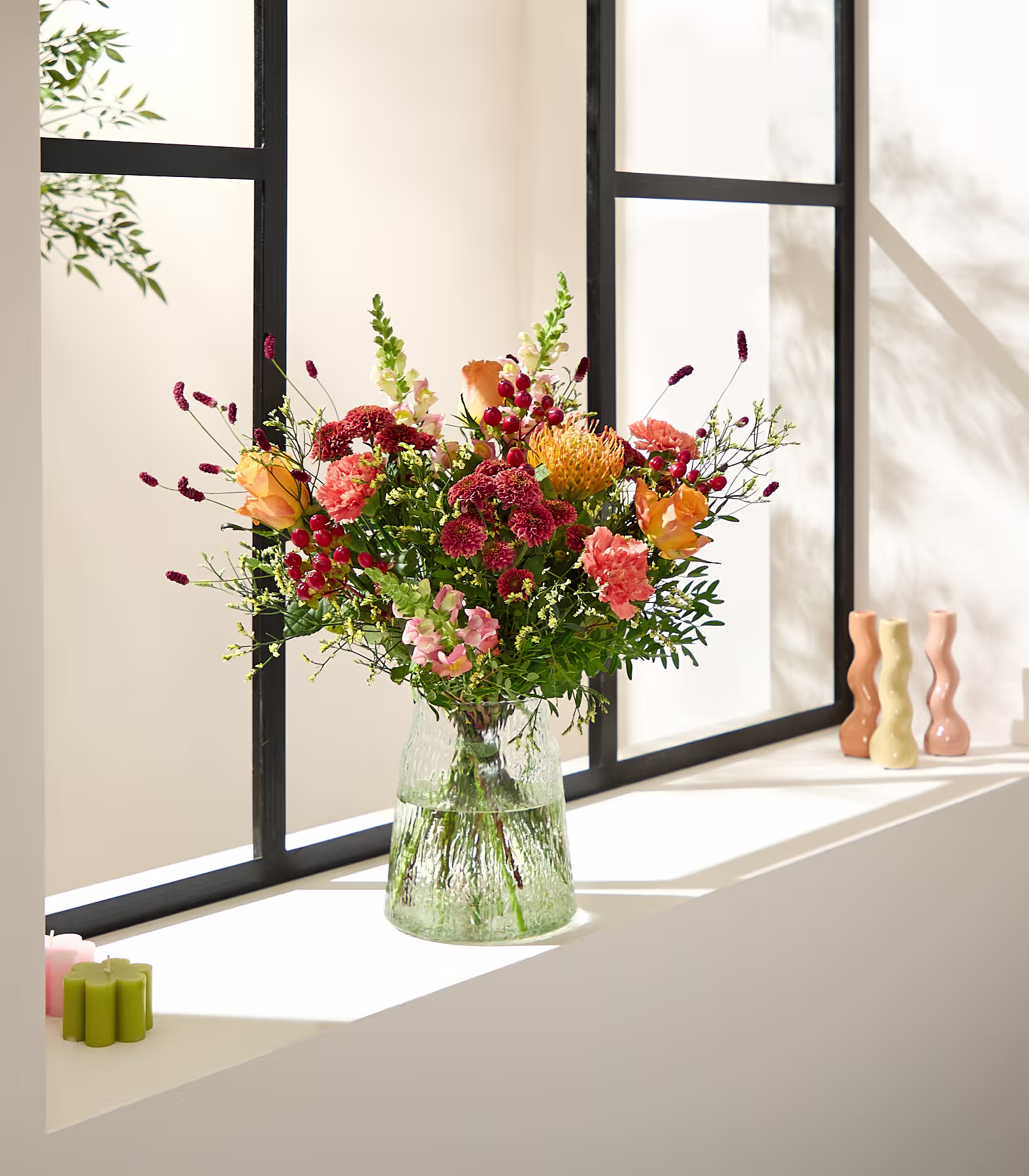 Auf einem Fensterbrett steht ein farbenfroher Strauß orangefarbener und roter Blumen in einer strukturierten Glasvase; im Hintergrund sind ein Fenster und dekorative Kerzen zu sehen. (Beschreibungstext KI-generiert)