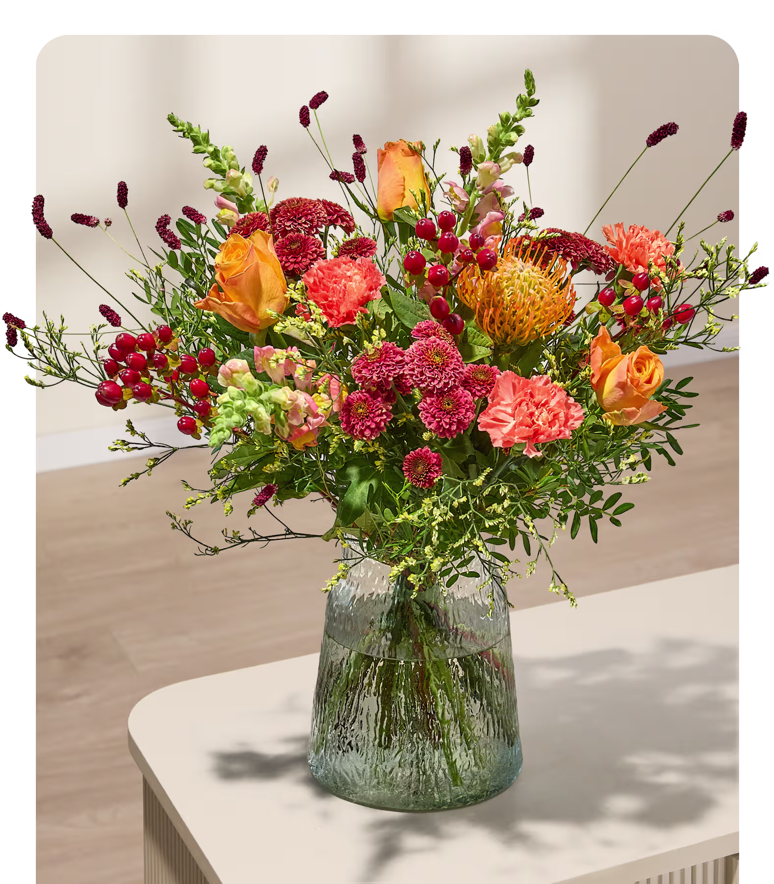 Ein leuchtender Strauß orangefarbener und rosafarbener Blumen mit roten Beeren steht in einer strukturierten Glasvase auf einem hellen Tisch, im Hintergrund sind ein Holzboden und eine Holzwand verschwommen zu sehen. (Beschreibungstext KI-generiert)