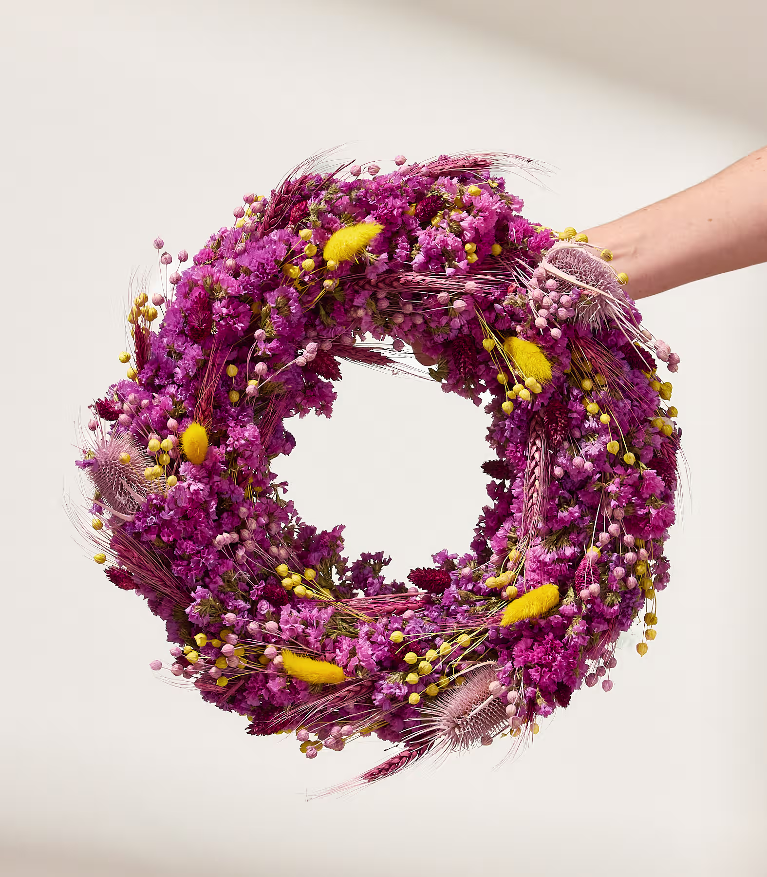 Eine Hand hält einen runden Kranz aus getrockneten lila und rosa Blüten mit gelben Akzenten vor einem schlichten, hellen Hintergrund. (Beschreibungstext KI-generiert)