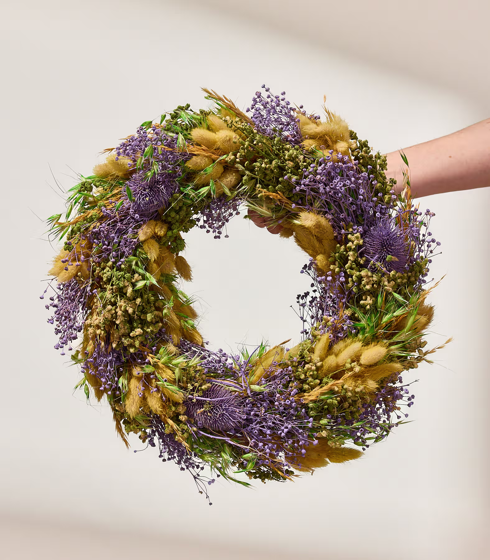 Eine Hand hält einen kreisförmigen Kranz aus getrockneten violetten Blüten und goldenen Gräsern vor einem sanft beleuchteten, neutralen Hintergrund. (Beschreibungstext KI-generiert)