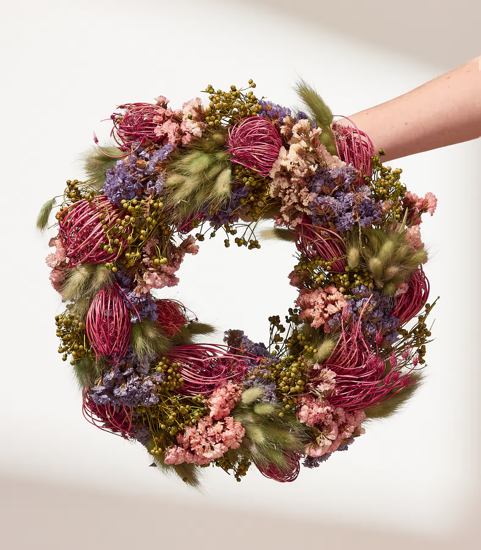 Eine Hand hält einen kreisförmigen Kranz aus getrockneten Blumen und Gräsern vor einem sanft beleuchteten, cremefarbenen Hintergrund. (Beschreibungstext KI-generiert)