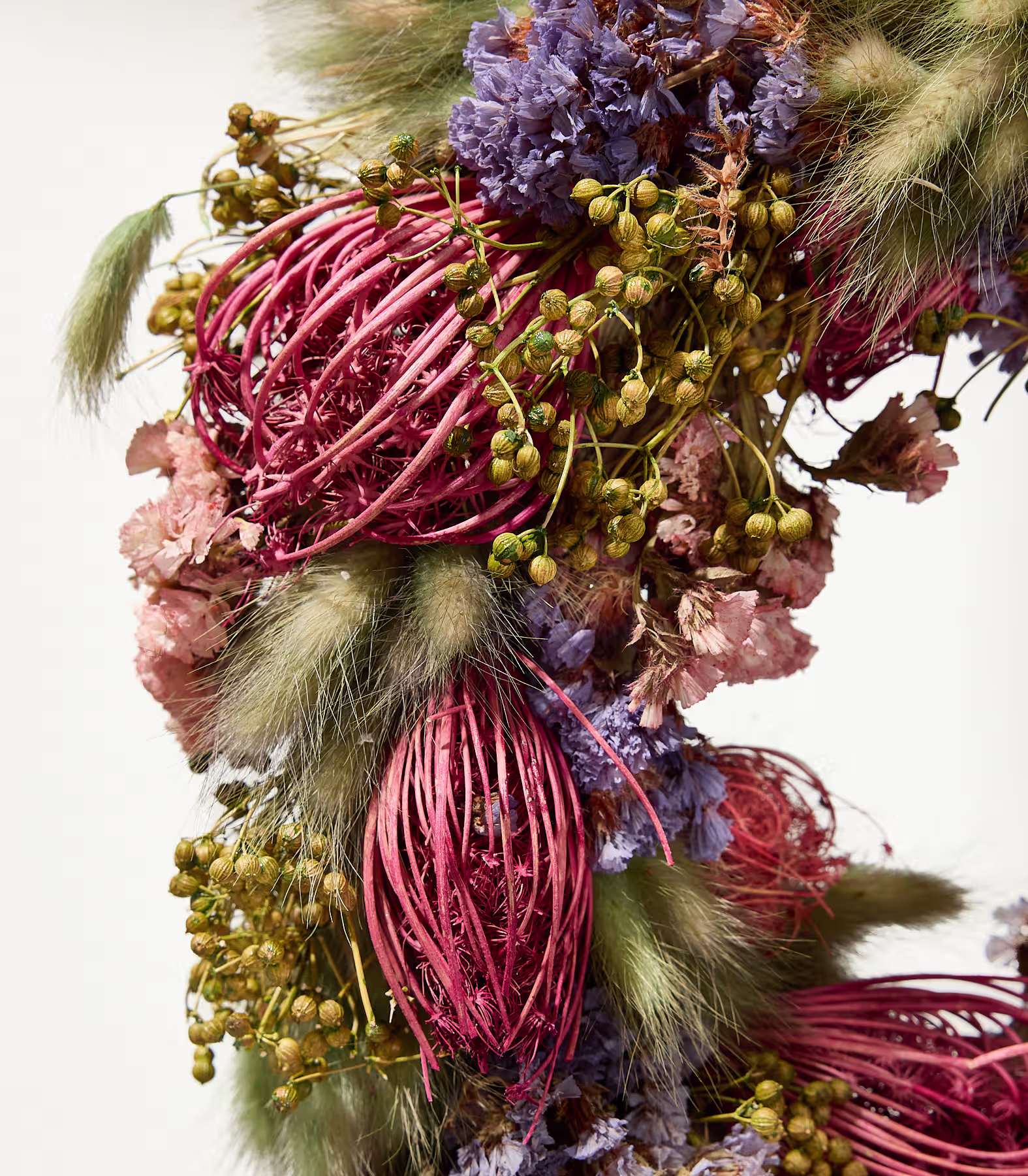 Eine Nahaufnahme eines Trockenblumenarrangements mit leuchtend pinken, violetten und grünen Elementen im Vordergrund vor einem schlichten weißen Hintergrund. (Beschreibungstext KI-generiert)