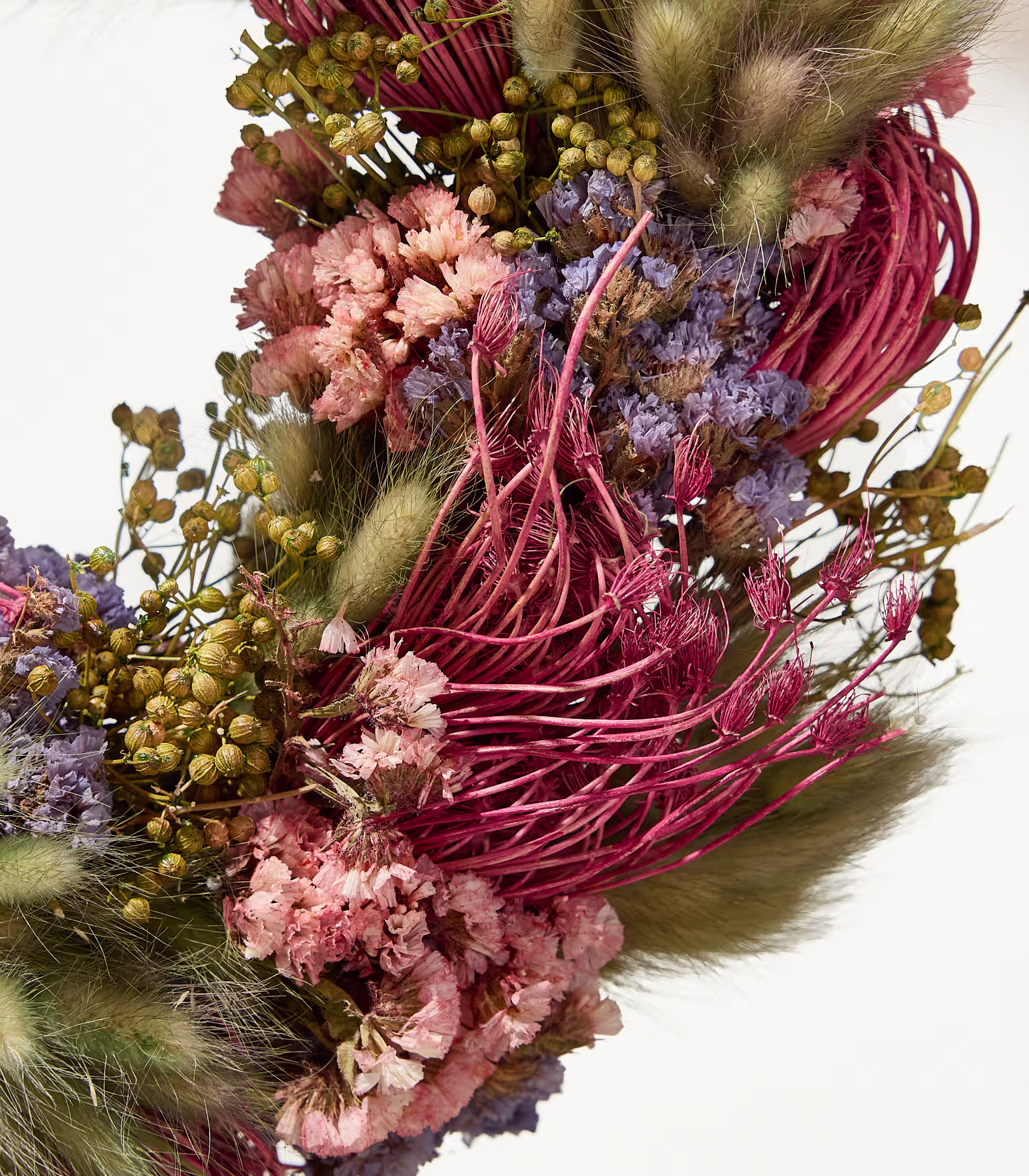 Eine Nahaufnahme eines Trockenblumenkranzes vor weißem Hintergrund zeigt im Vordergrund eine lebhafte Mischung aus rosa, violetten und grünen Pflanzen. (Beschreibungstext KI-generiert)