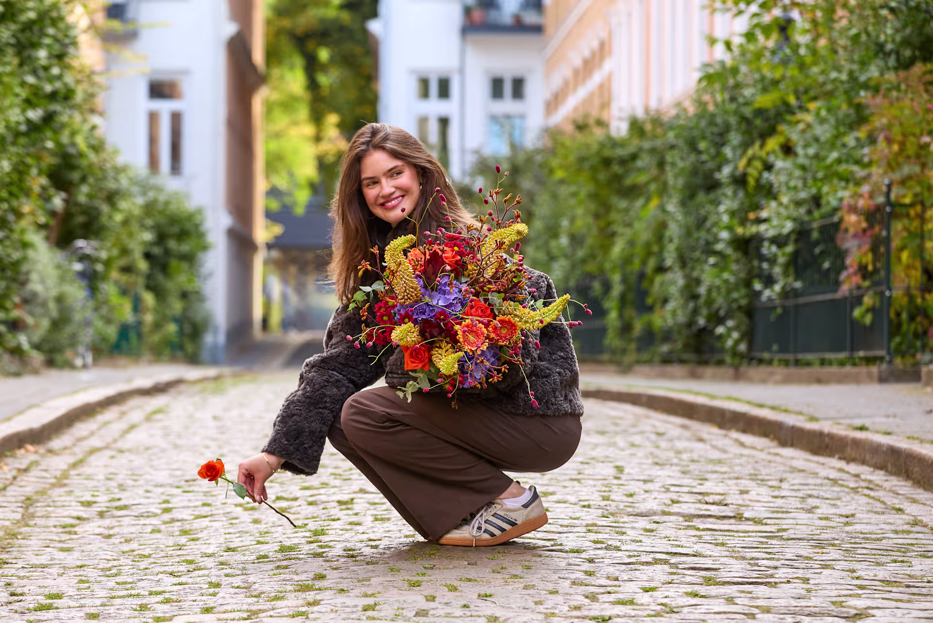 Eine lächelnde Frau hockt auf einem Kopfsteinpflaster, hält einen farbenprächtigen Blumenstrauß in der Hand; im Hintergrund erheben sich Gebäude und Grünanlagen. (Beschreibungstext KI-generiert)