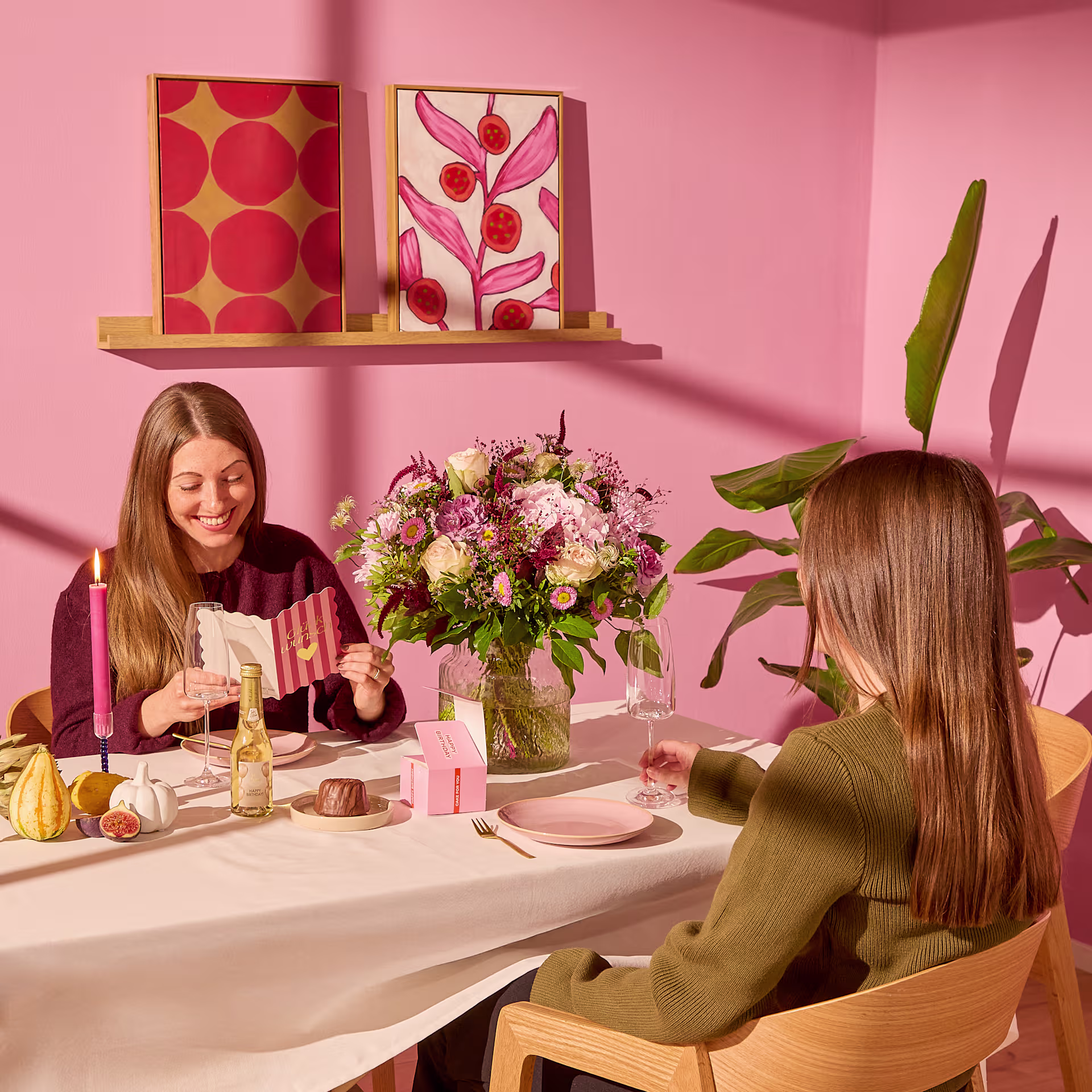 Zwei Frauen sitzen an einem mit Blumenarrangements und Desserts gedeckten Tisch; im Hintergrund sind rosa Wände und Kunstwerke zu sehen. (Beschreibungstext KI-generiert)