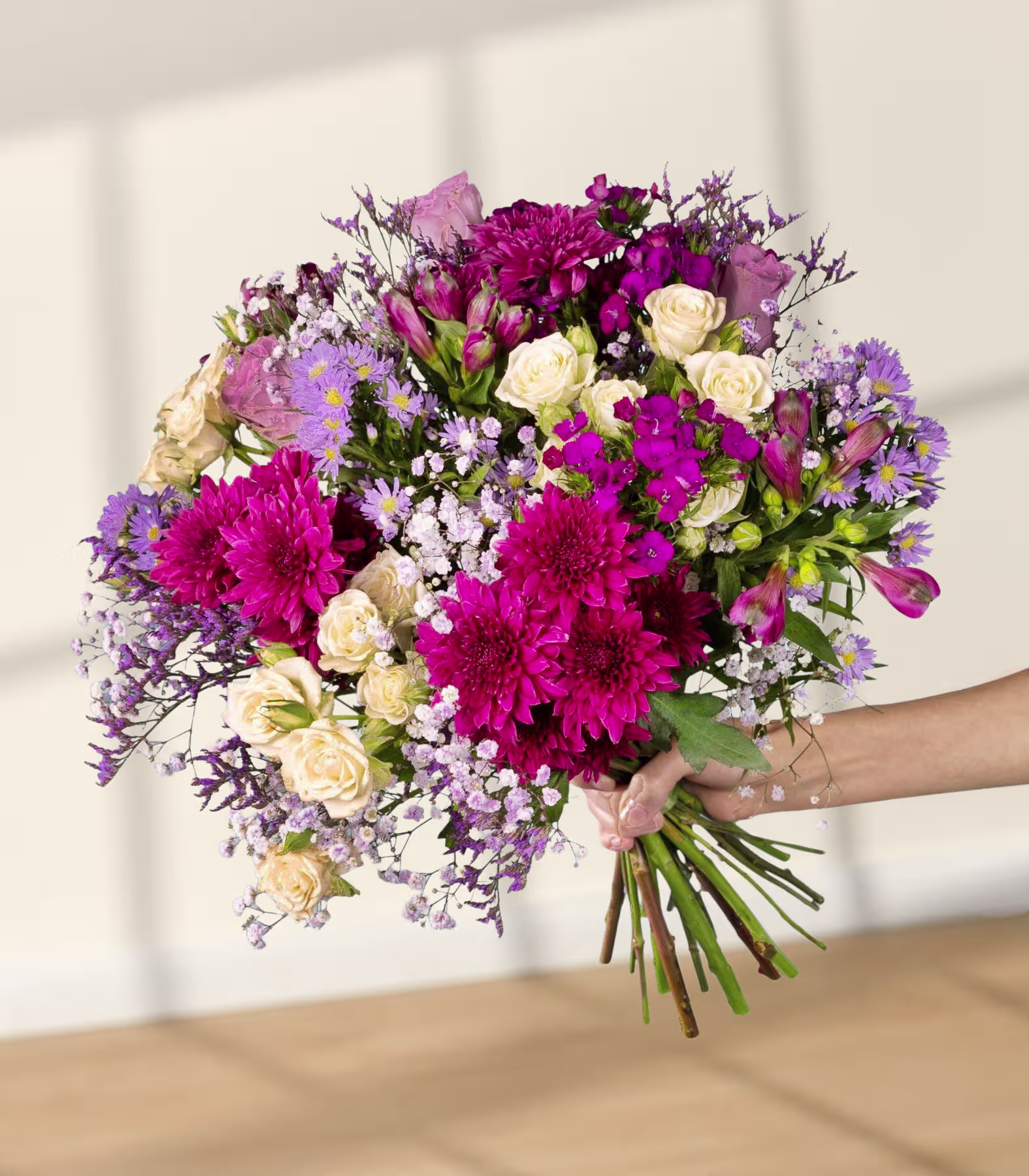 Ein lebhafter Strauß aus verschiedenen bunten Blumen, darunter rosa und violette Chrysanthemen, weiße Rosen und zarte violette und weiße Blüten, die in den Händen einer Person vor einem schlichten weißen Hintergrund gehalten werden. (Beschreibungstext KI-generiert)