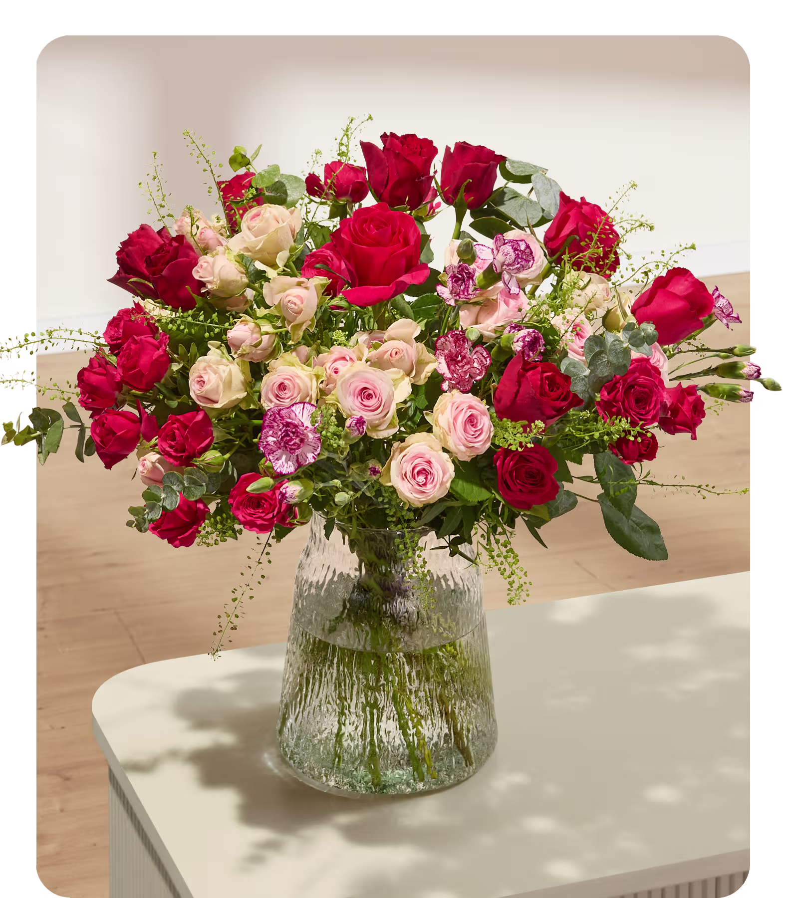 Ein farbenprächtiger Strauß roter und rosa Rosen füllt eine strukturierte Glasvase auf einem hellen Tisch, im Hintergrund ist eine sanft beleuchtete Wand zu sehen. (Beschreibungstext KI-generiert)