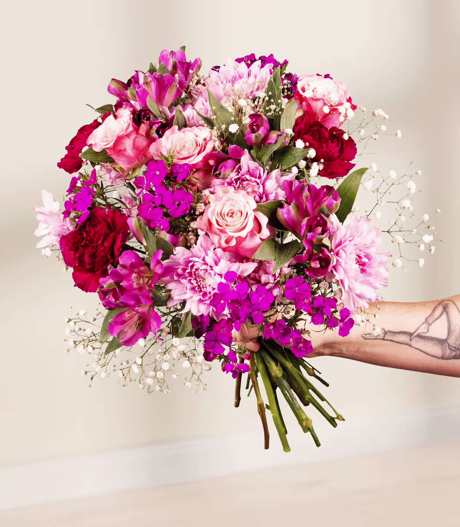 Ein farbenfroher Blumenstrauß aus rosa, roten und violetten Blüten, darunter Rosen, Nelken und Schleierkraut, in der tätowierten Hand einer Person vor einem schlichten blauen Hintergrund. (Beschreibungstext KI-generiert)