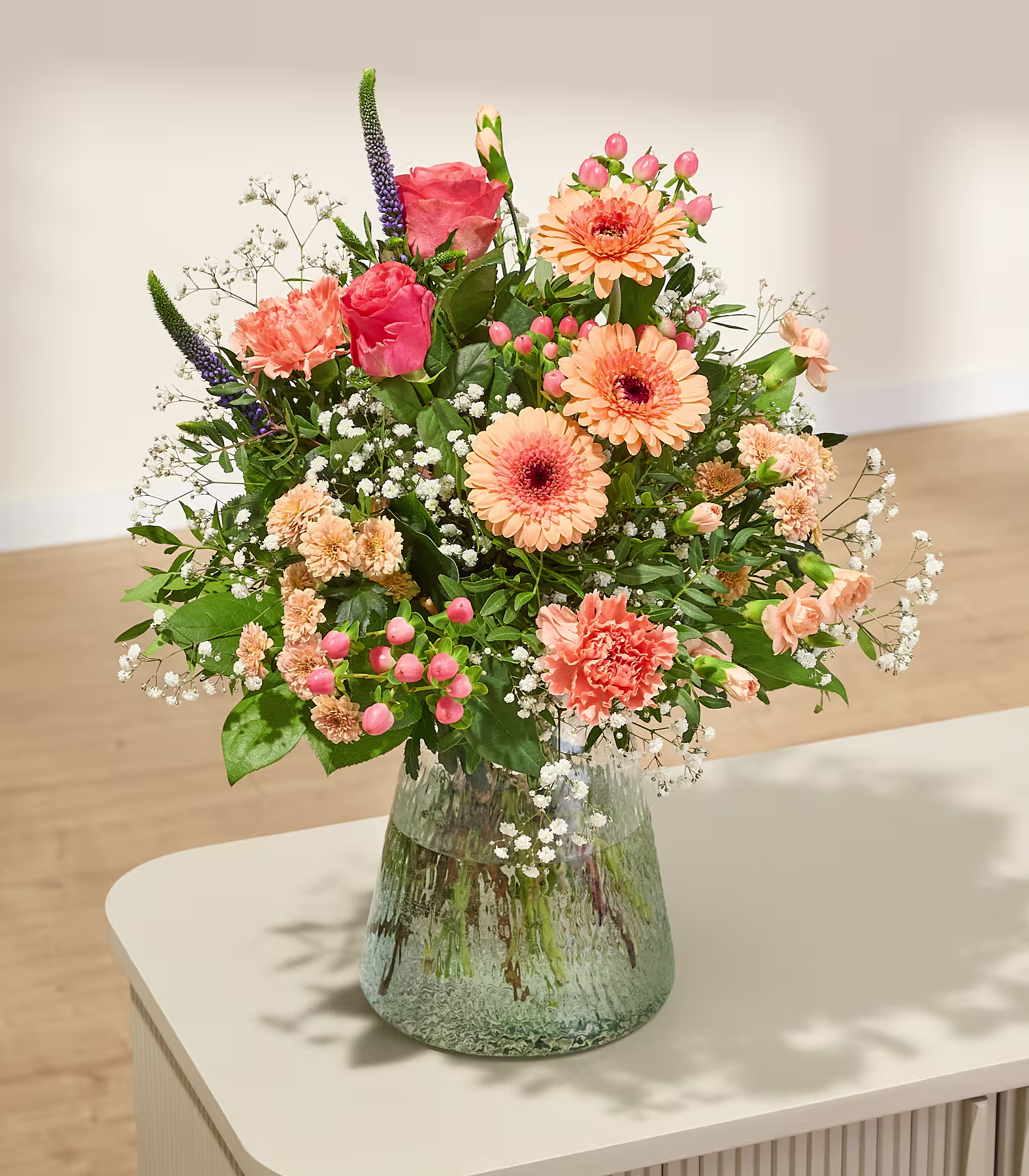 Ein leuchtender Strauß aus orangefarbenen und rosafarbenen Blumen mit zartem weißen Schleierkraut füllt eine Glasvase auf einer hellen Oberfläche vor einem sanft beleuchteten Hintergrund mit gefleckten Schatten. (Beschreibungstext KI-generiert)