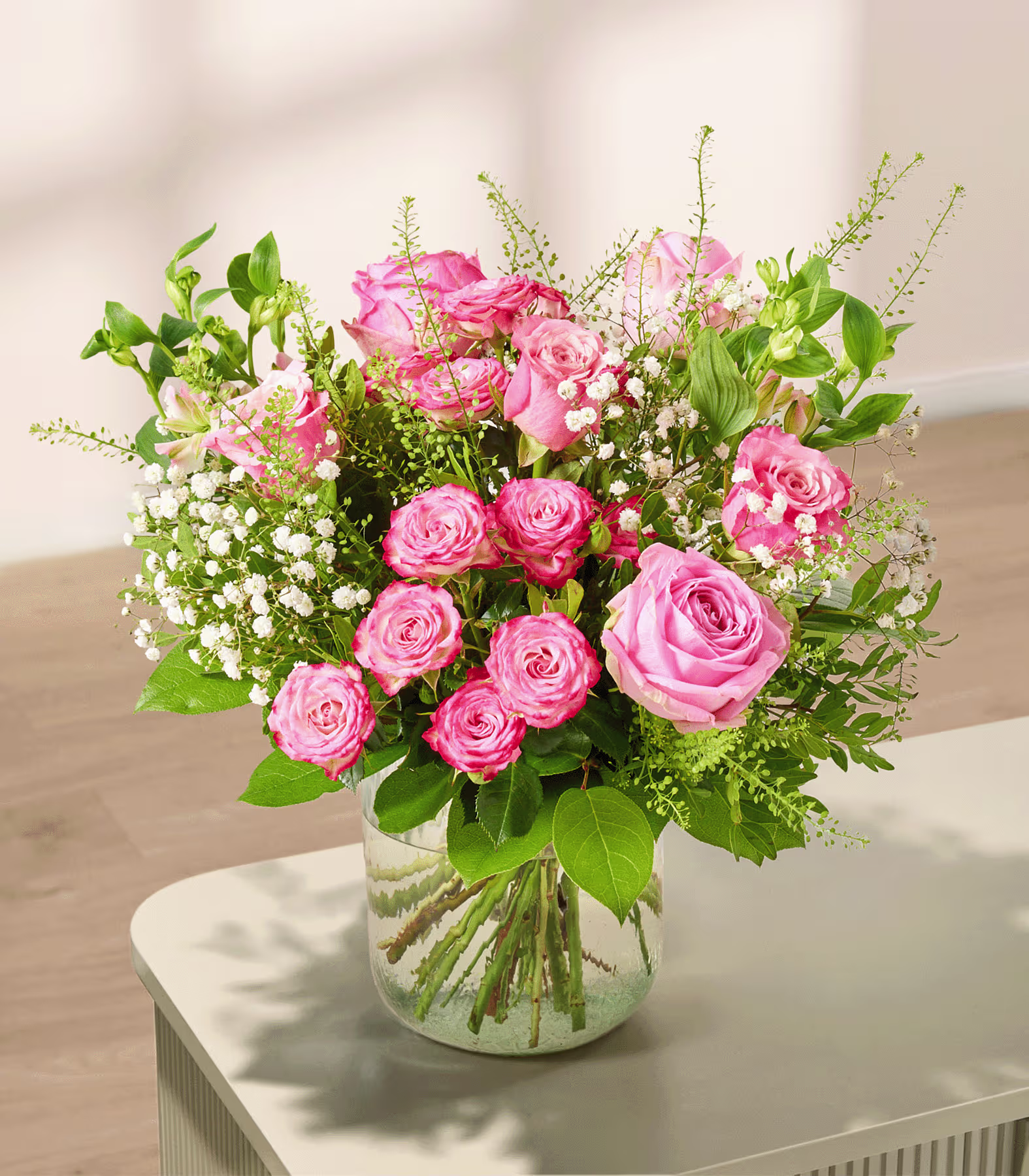 Ein Strauß rosa Rosen und Schleierkraut steht in einer Glasvase auf einem hellen Tisch, im Hintergrund ist eine sanft beleuchtete Wand zu sehen. (Beschreibungstext KI-generiert)