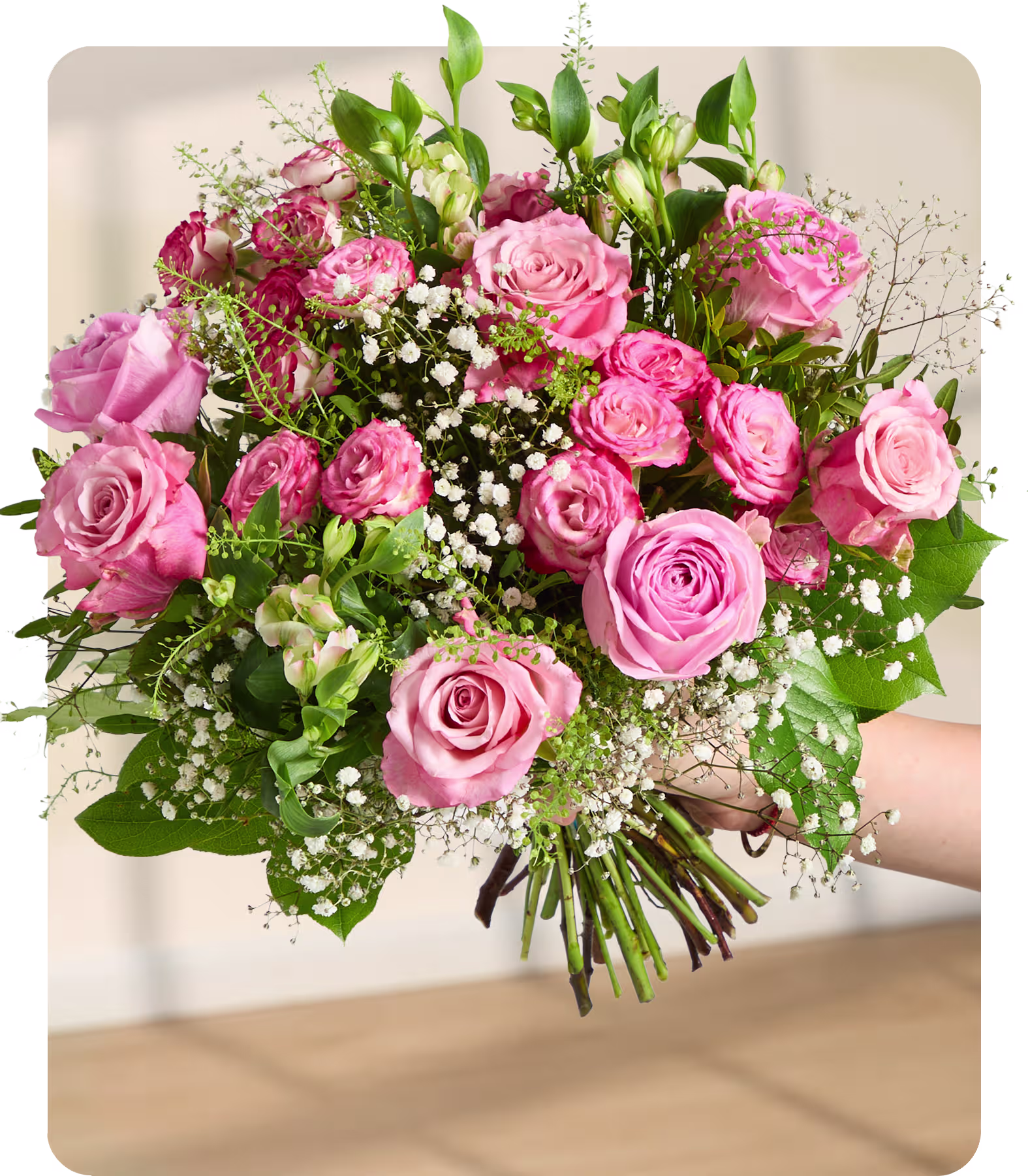 Eine Hand hält einen üppigen Strauß rosa Rosen und Schleierkraut vor einem sanft verschwommenen Hintergrund aus hellen Wänden und einer Holzoberfläche. (Beschreibungstext KI-generiert)
