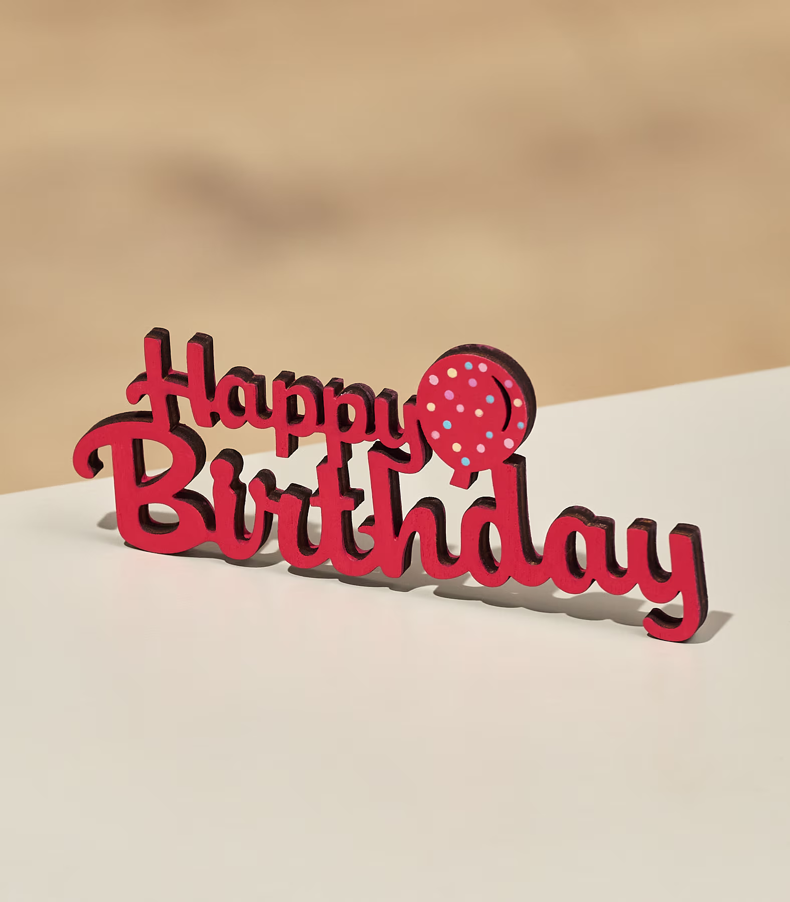 Ein rotes Schild mit der Aufschrift „Happy Birthday“ und einem gepunkteten Luftballon steht auf einer hellen Fläche vor einem schlichten Hintergrund. (Beschreibungstext KI-generiert)