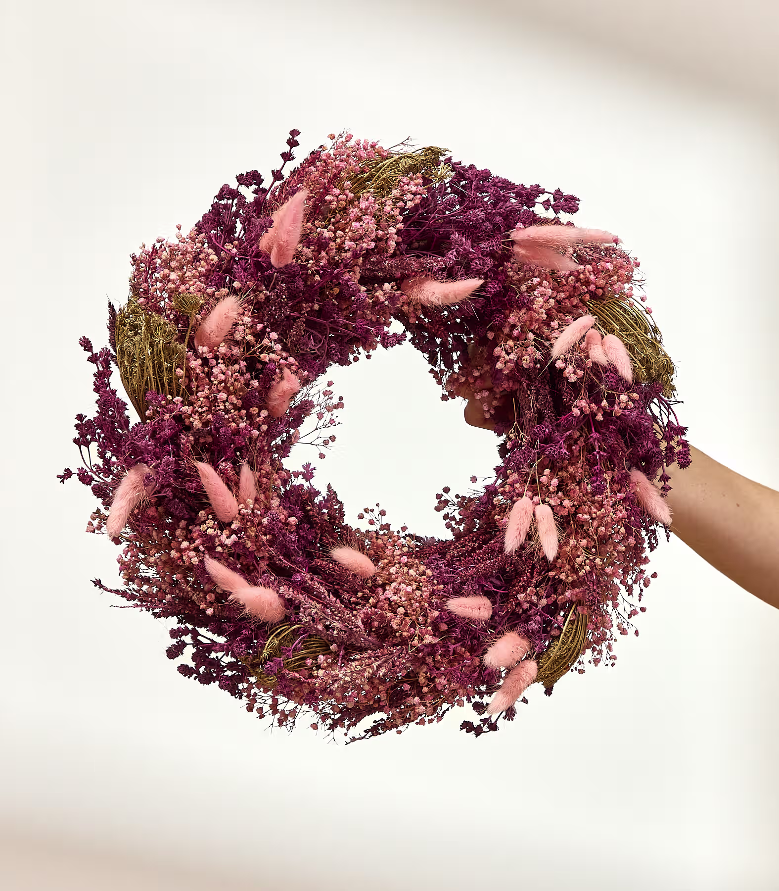 Eine Hand hält einen üppigen Kranz aus rosa und violetten Trockenblumen vor einem schlichten weißen Hintergrund. (Beschreibungstext KI-generiert)