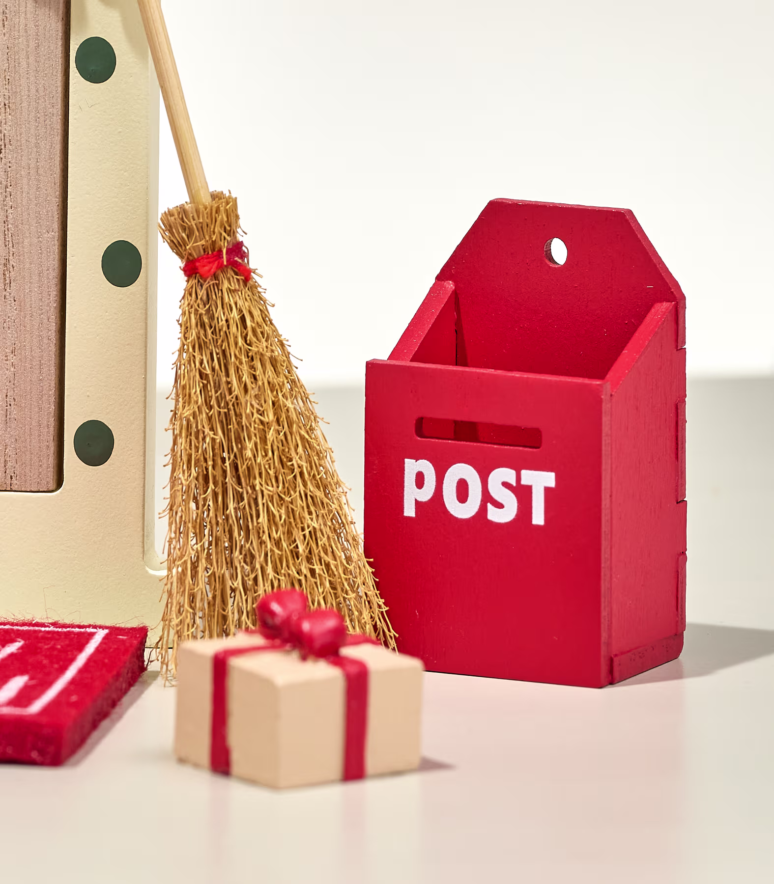 Im Vordergrund stehen eine kleine Geschenkbox und ein Strohbesen neben einem roten Miniaturbriefkasten vor einem schlichten weißen Hintergrund. (Beschreibungstext KI-generiert)