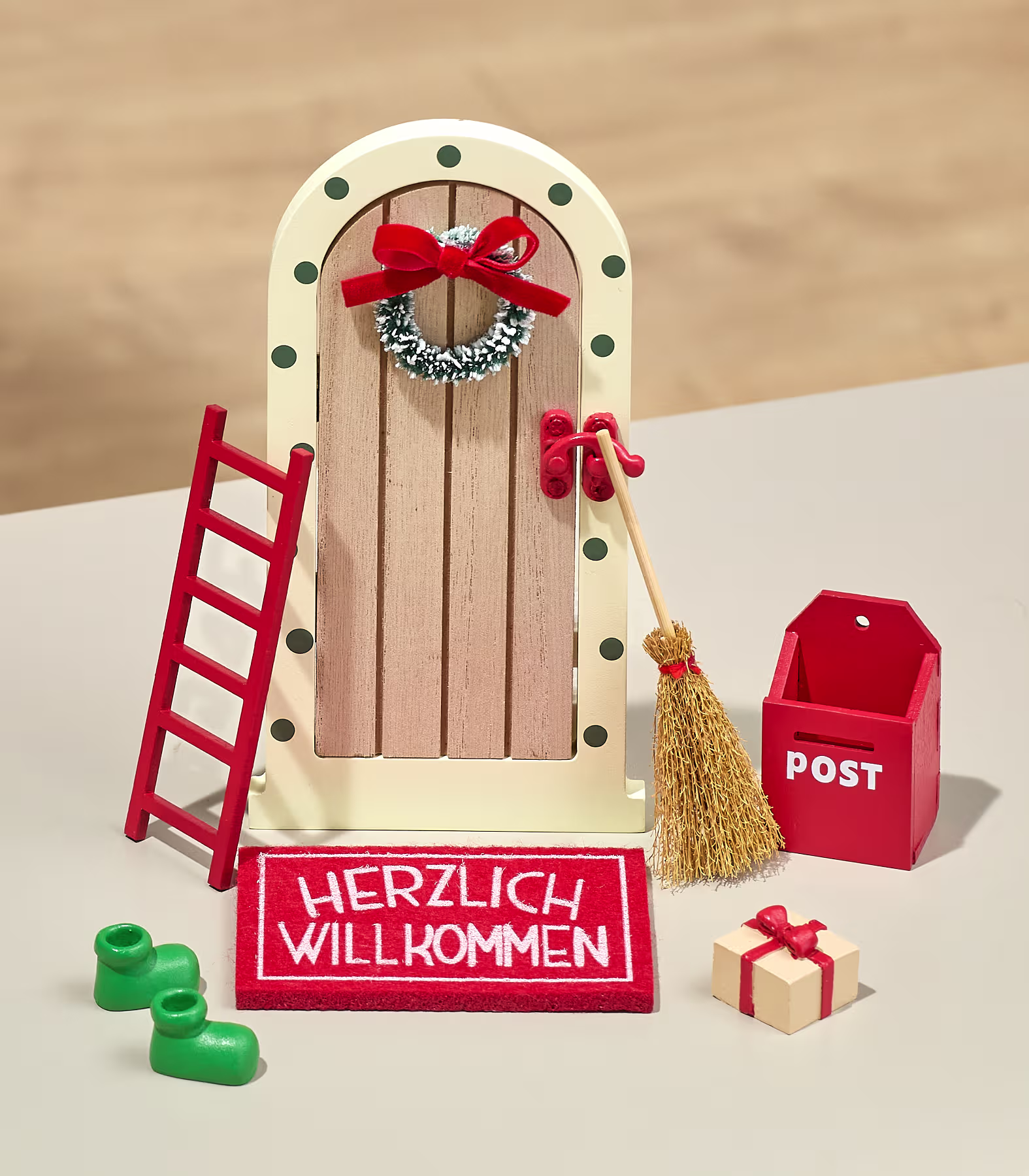 Eine Miniatur-Feentür, verziert mit einem Kranz, steht umgeben von einer roten Leiter, einem Besen, einem Briefkasten und einer Fußmatte vor einem neutralen, verschwommenen Hintergrund. (Beschreibungstext KI-generiert)