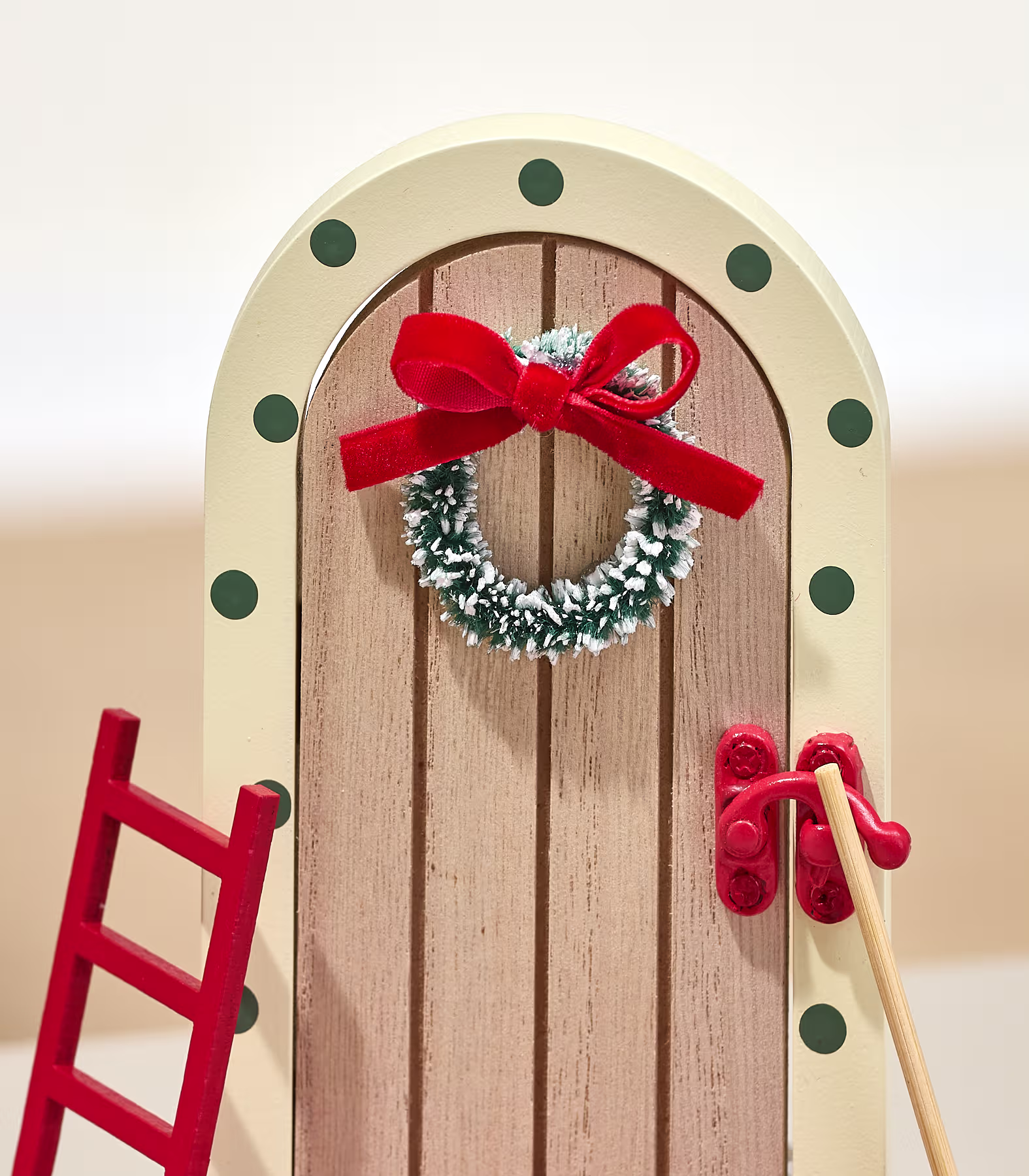 Eine Miniatur-Holztür, verziert mit einem schneebedeckten Kranz und einer roten Schleife, steht neben einer kleinen roten Leiter vor einem schlichten, hellen Hintergrund. (Beschreibungstext KI-generiert)