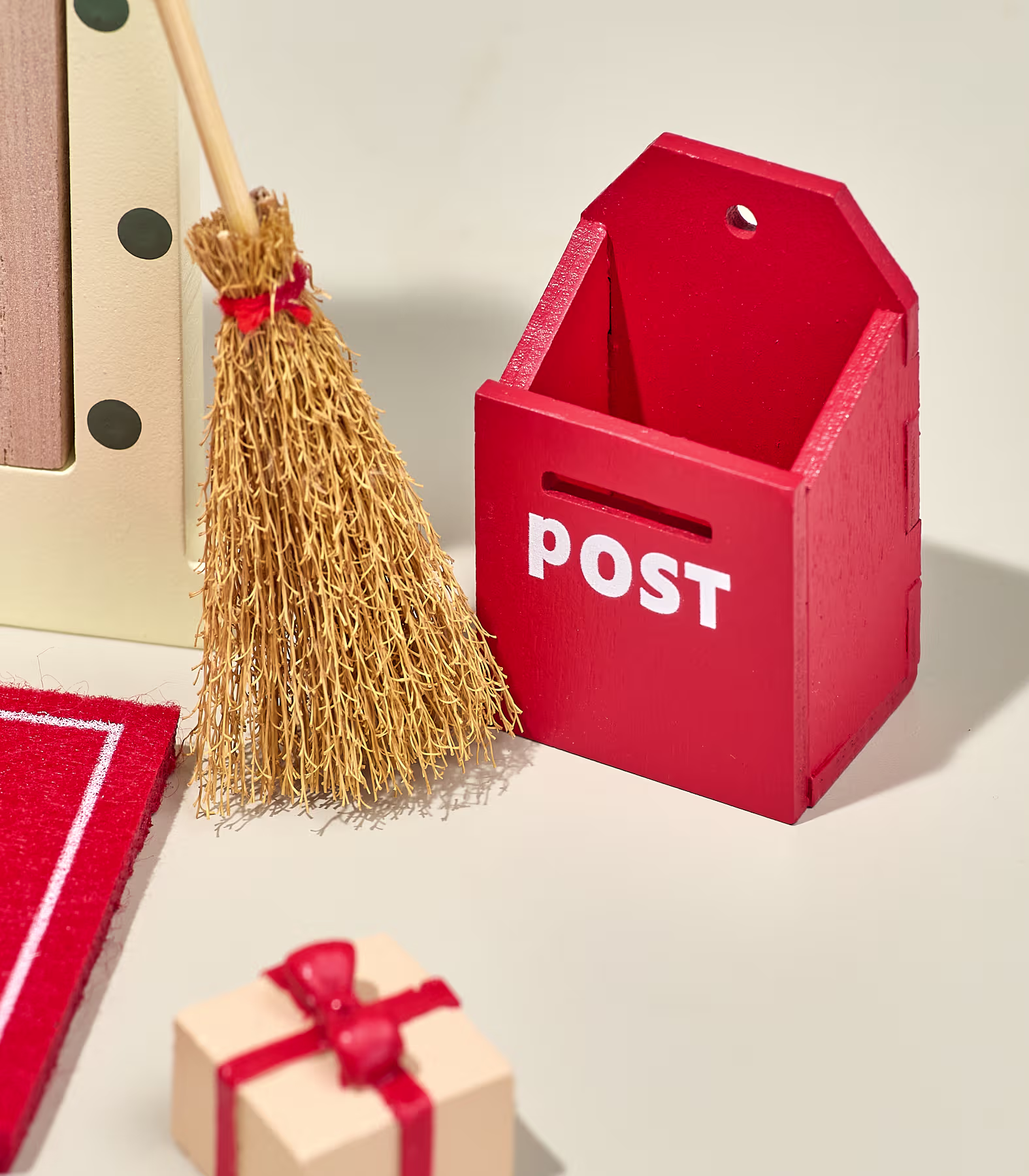 Im Vordergrund stehen ein Miniaturbesen und ein kleines, verpacktes Geschenk neben einem roten Holzbriefkasten vor einem schlichten, hellen Hintergrund. (Beschreibungstext KI-generiert)