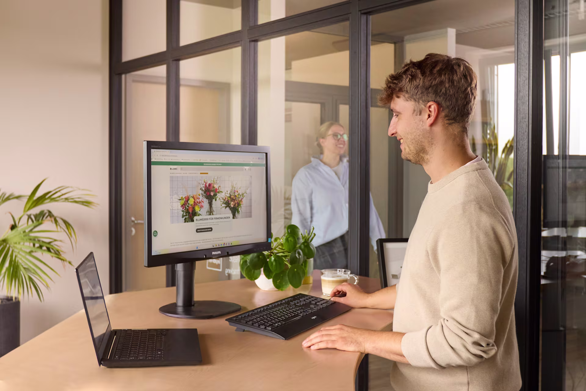 Ein Mann in einem beigen Pullover arbeitet an einem Schreibtisch mit einem Monitor und einem Laptop im Vordergrund, während im Hintergrund durch eine Glastrennwand eine Frau zu sehen ist. (Beschreibungstext KI-generiert)